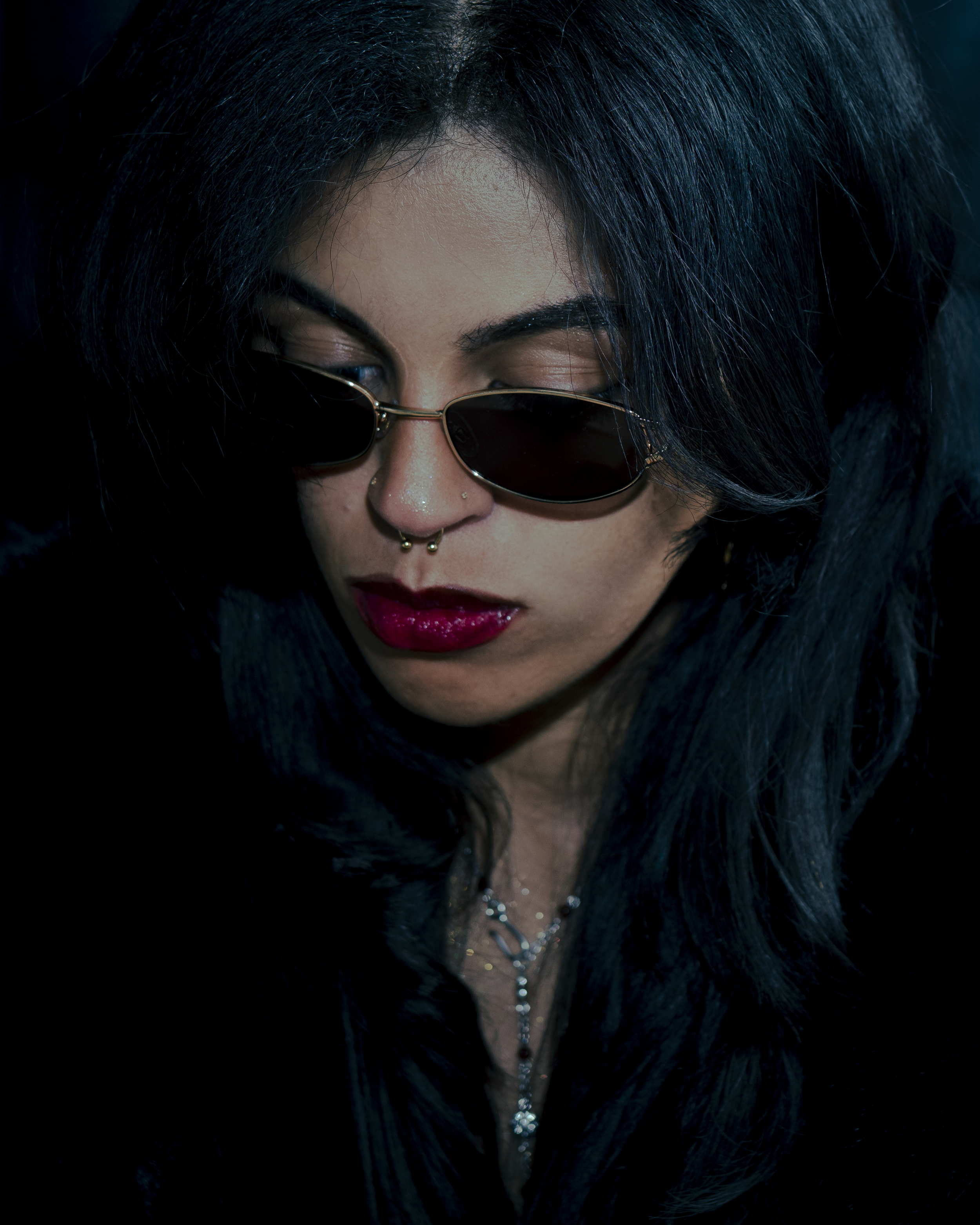 A woman with long dark hair, wearing sunglasses, red lipstick, and jewelry, with a dark background.
