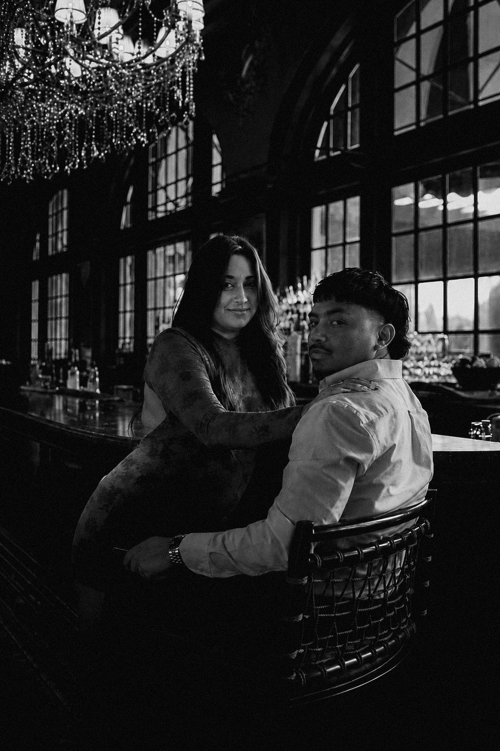 A black-and-white photo of a man and woman sitting at a bar inside a stylish restaurant or lounge. The woman has long hair and is smiling softly, wearing a long-sleeve top. The man, with short hair, is looking toward the camera, wearing a button-up shirt. Large windows and a chandelier with hanging crystals are visible in the background.