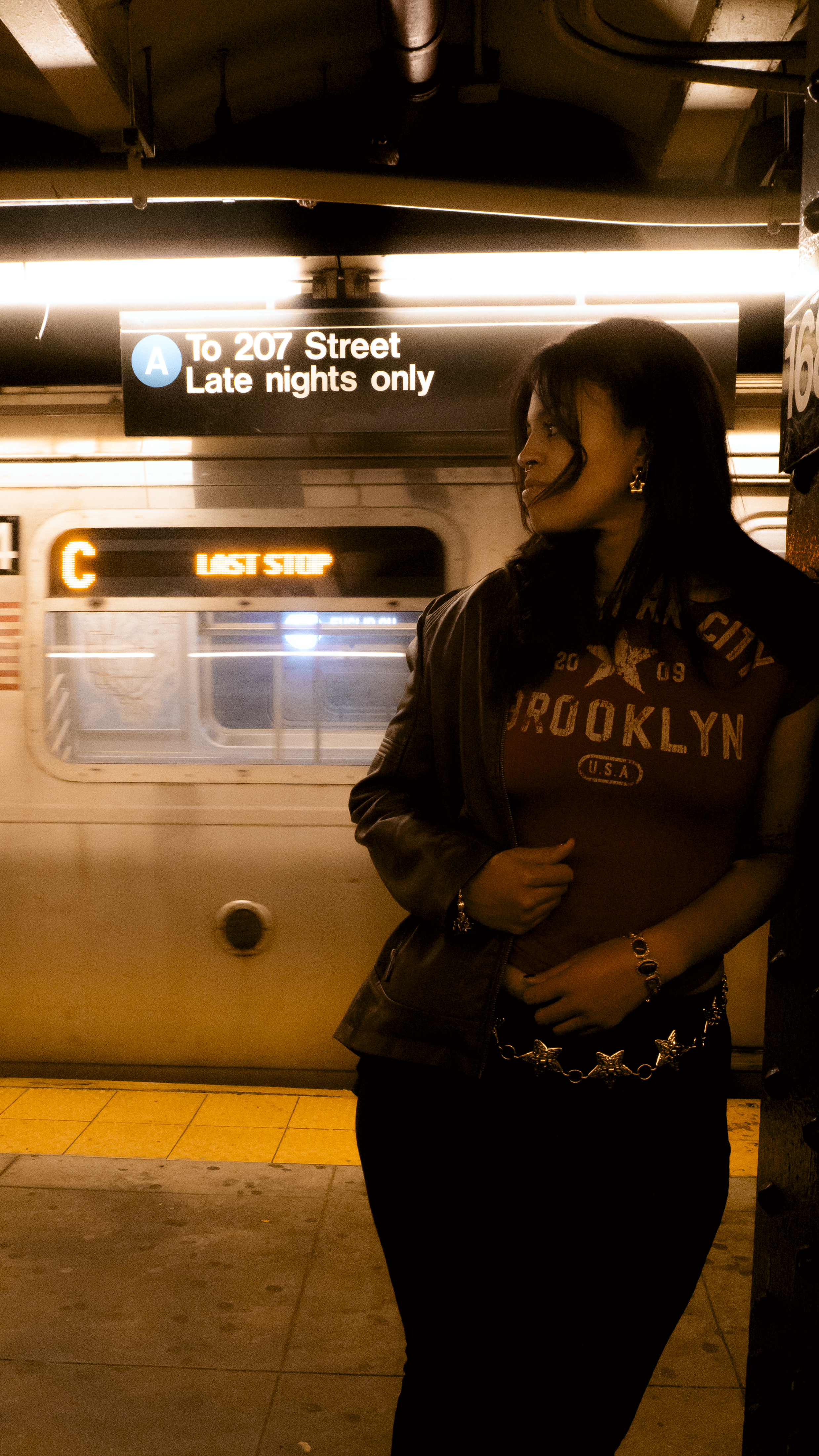 A woman standing on a subway platform at night with a train approaching. She is wearing a dark jacket and a red shirt that says Brooklyn U.S.A. and has gold jewelry. The train's destination signs show 'C' and 'LAST STOP.'