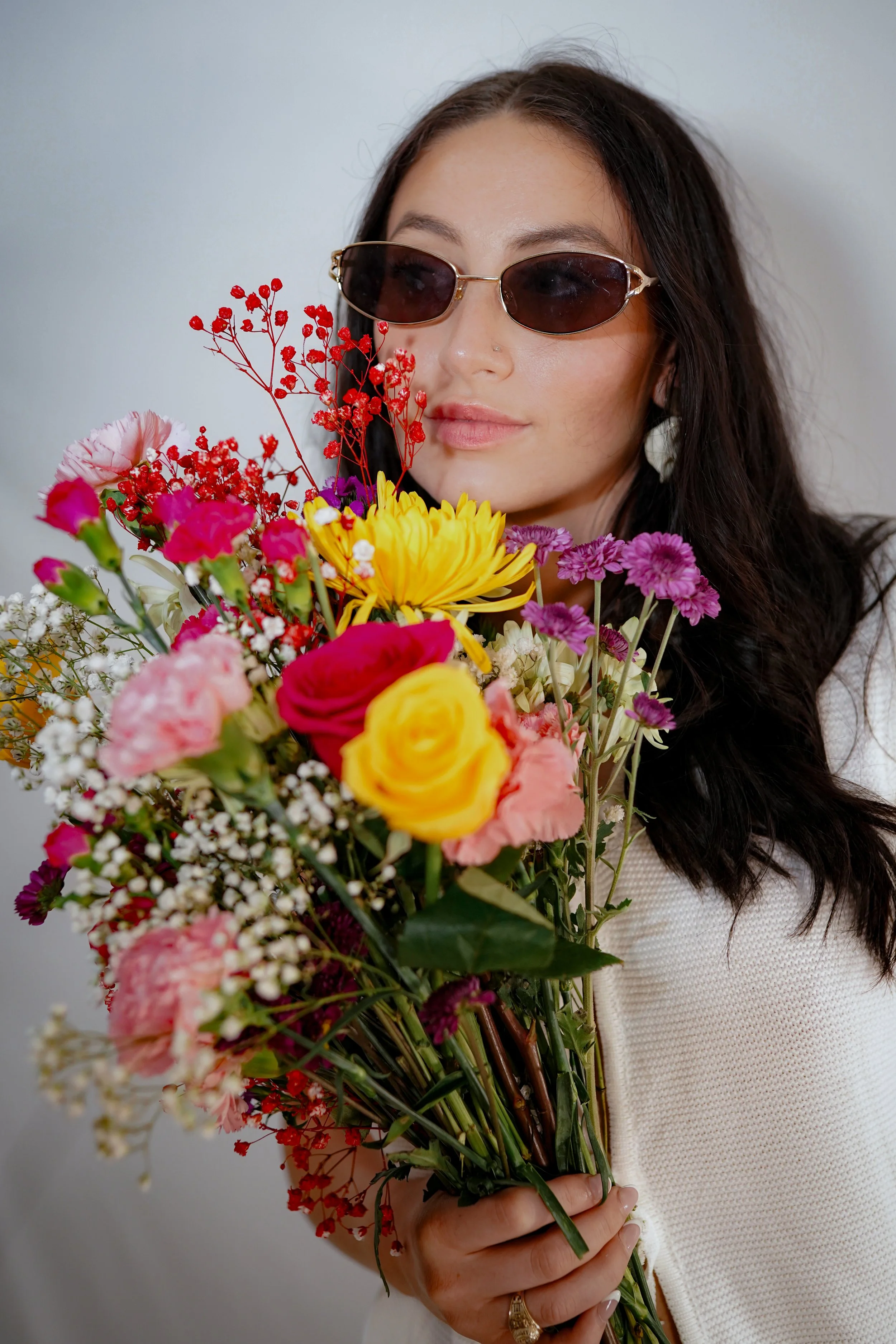 A woman wearing sunglasses holding a large colorful bouquet of flowers, including yellow, pink, purple, and white flowers against a plain gray background.