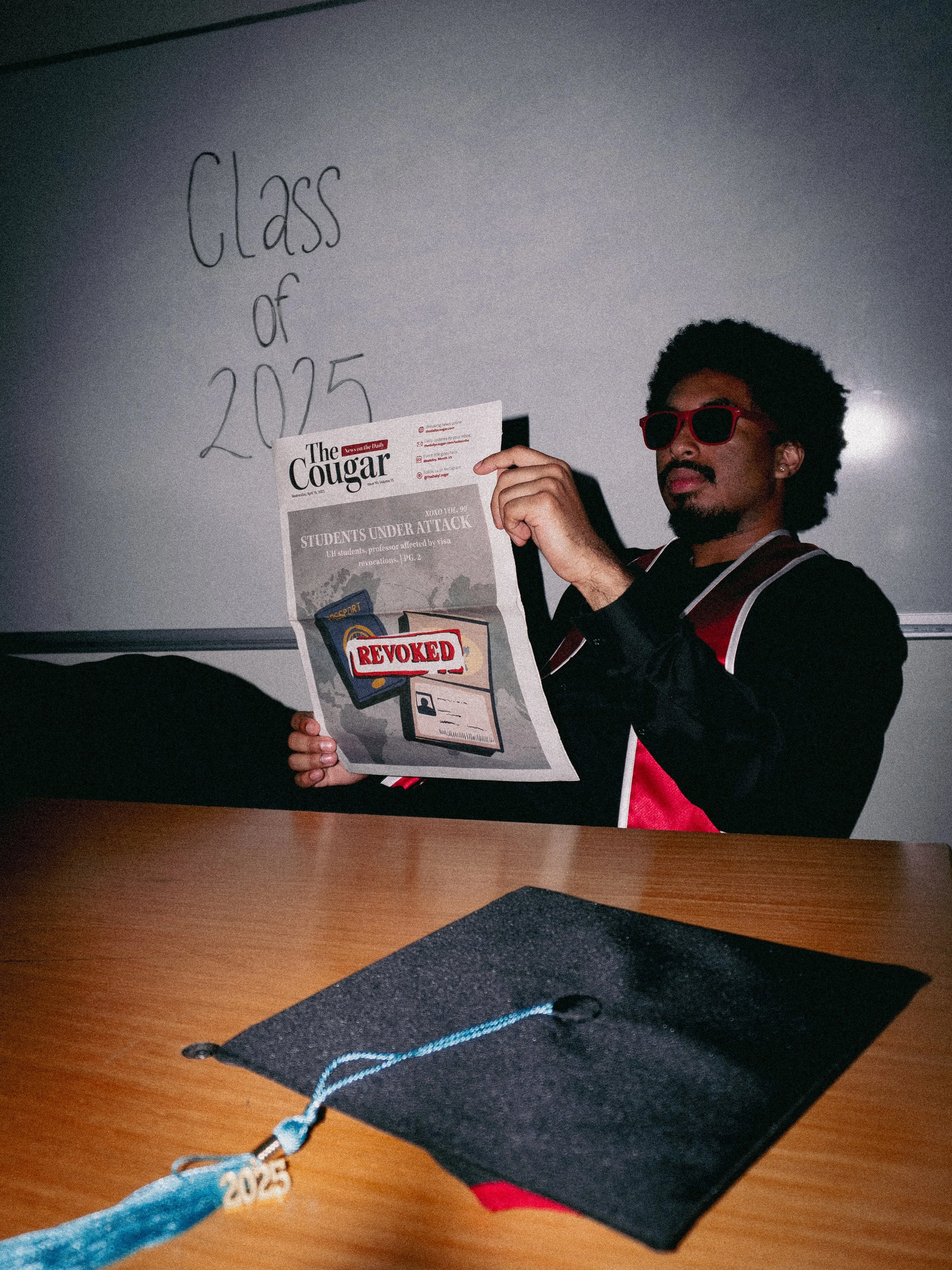 A person with an afro hairstyle and sunglasses holding a newspaper in a classroom setting. The whiteboard behind displays 'Class of 2025' written on it. A graduation cap and a blue tassel with '2025' are on a wooden table in the foreground.