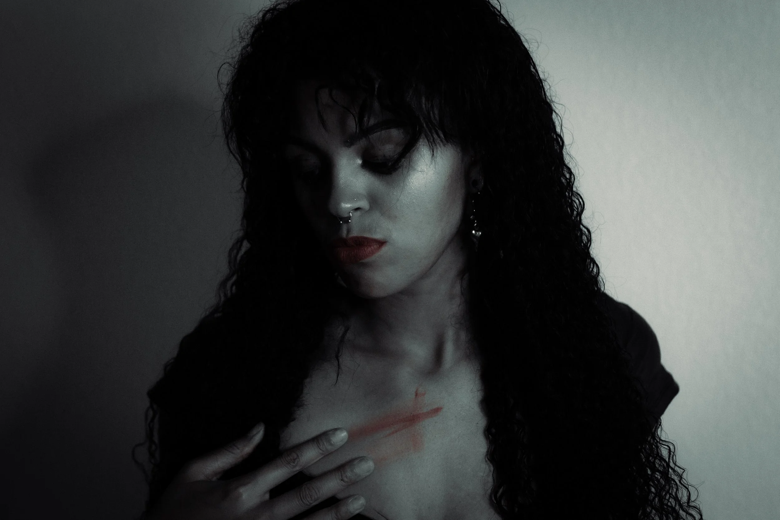 A woman with curly black hair and red lipstick has her hand on her chest, with red lipstick smudged across her collarbone. The image is dark and moody, with a plain wall background.