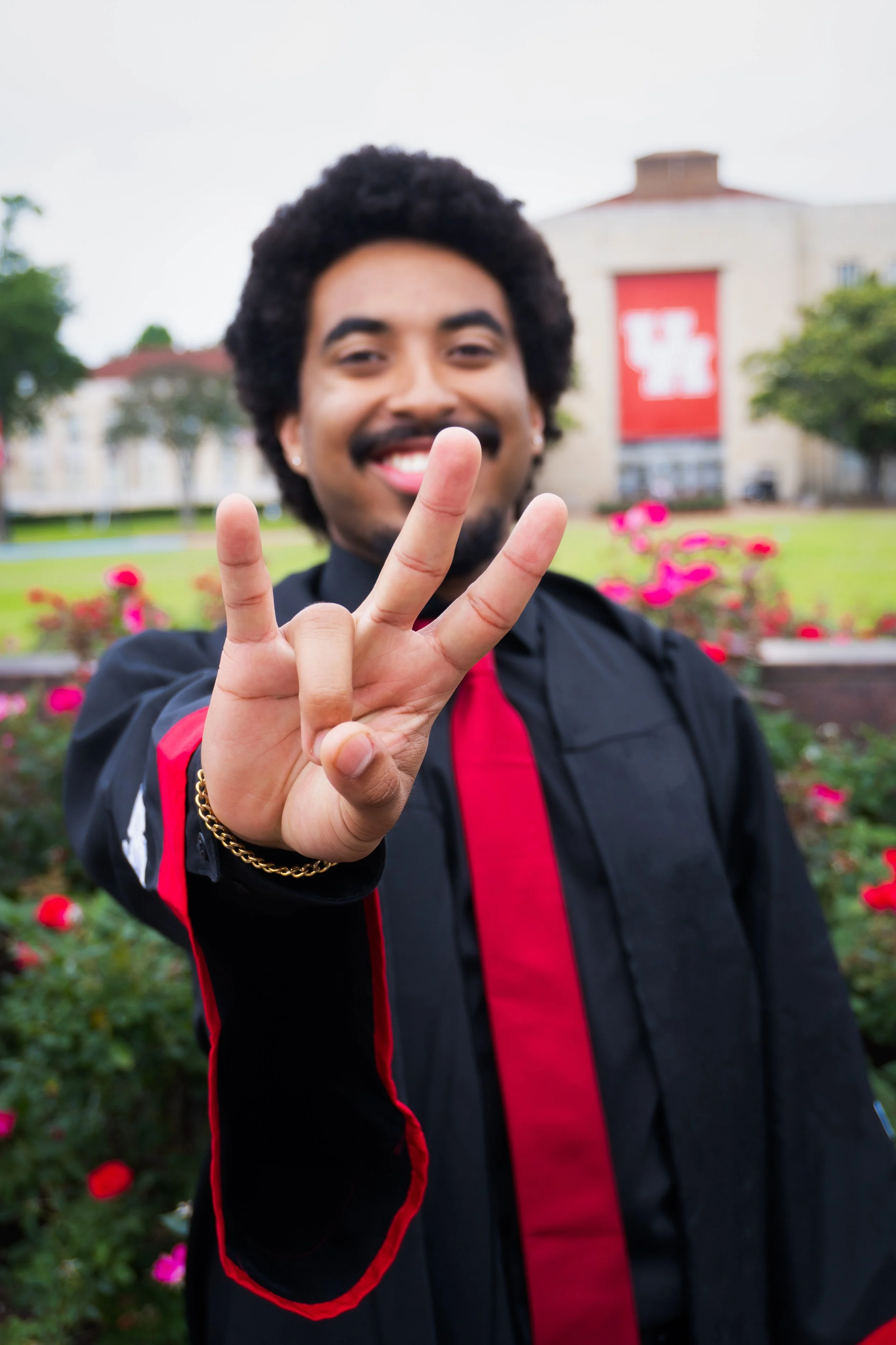 A young man with curly hair and a mustache smiling and making a peace sign with his fingers in the foreground. There are pink flowers and greenery behind him, and a building with a large red banner.