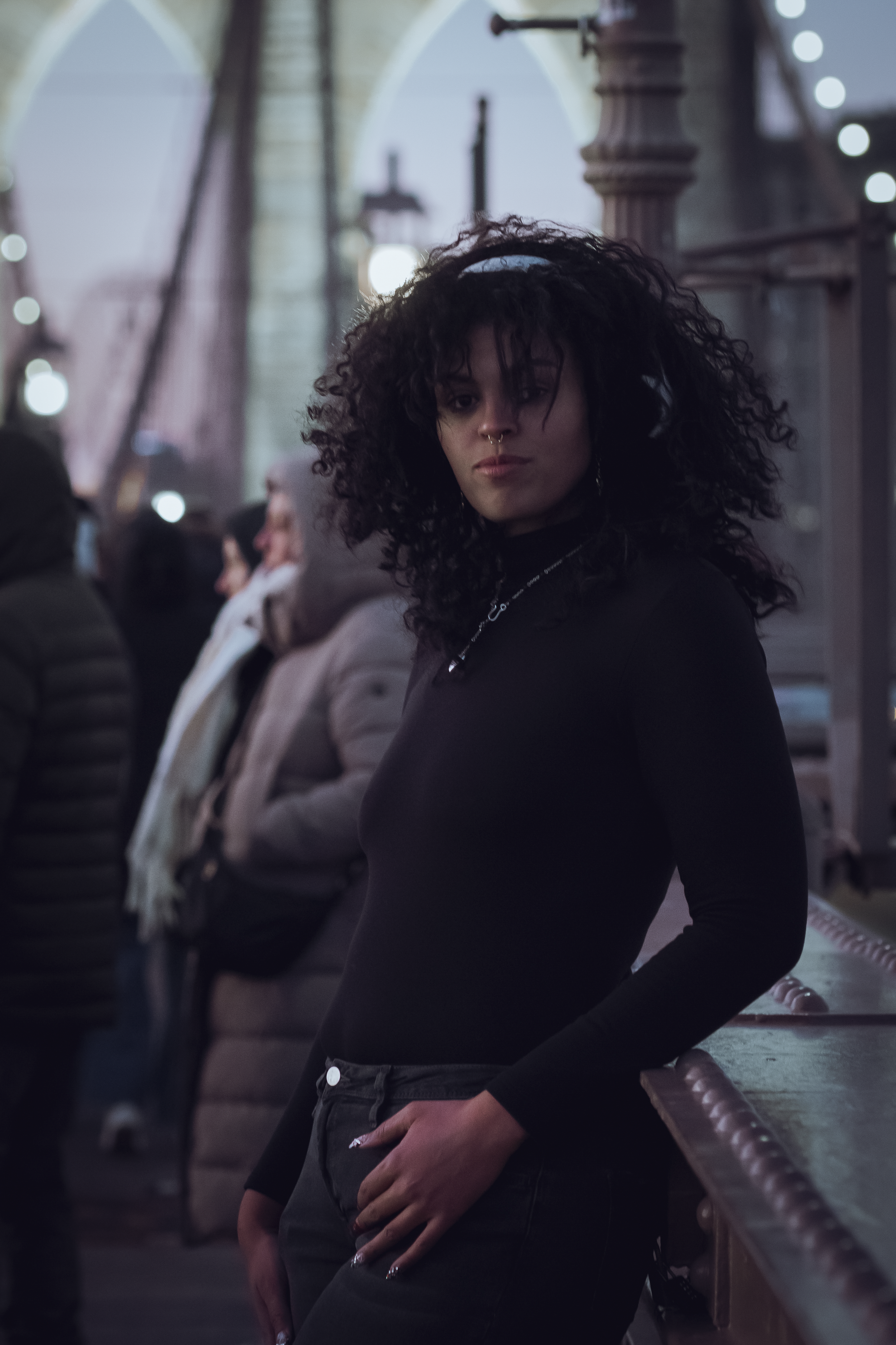 A woman with curly black hair, wearing a black long-sleeve shirt and dark pants, leaning against a railing on a bridge or walkway. The background is blurred, with a few people and city lights visible.