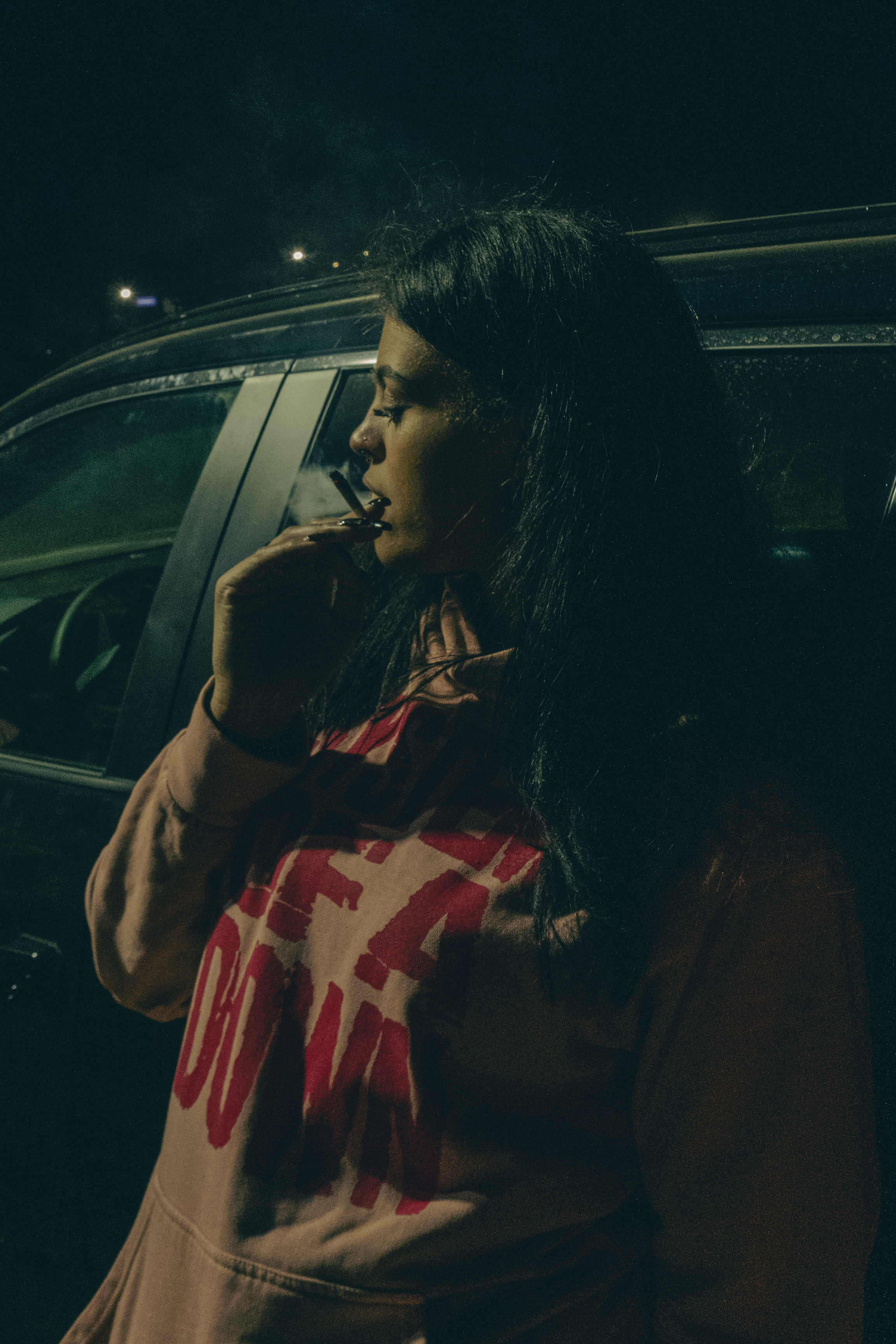 A woman with long dark hair, closed eyes, and a nose ring, smoking a cigarette inside a car at night.