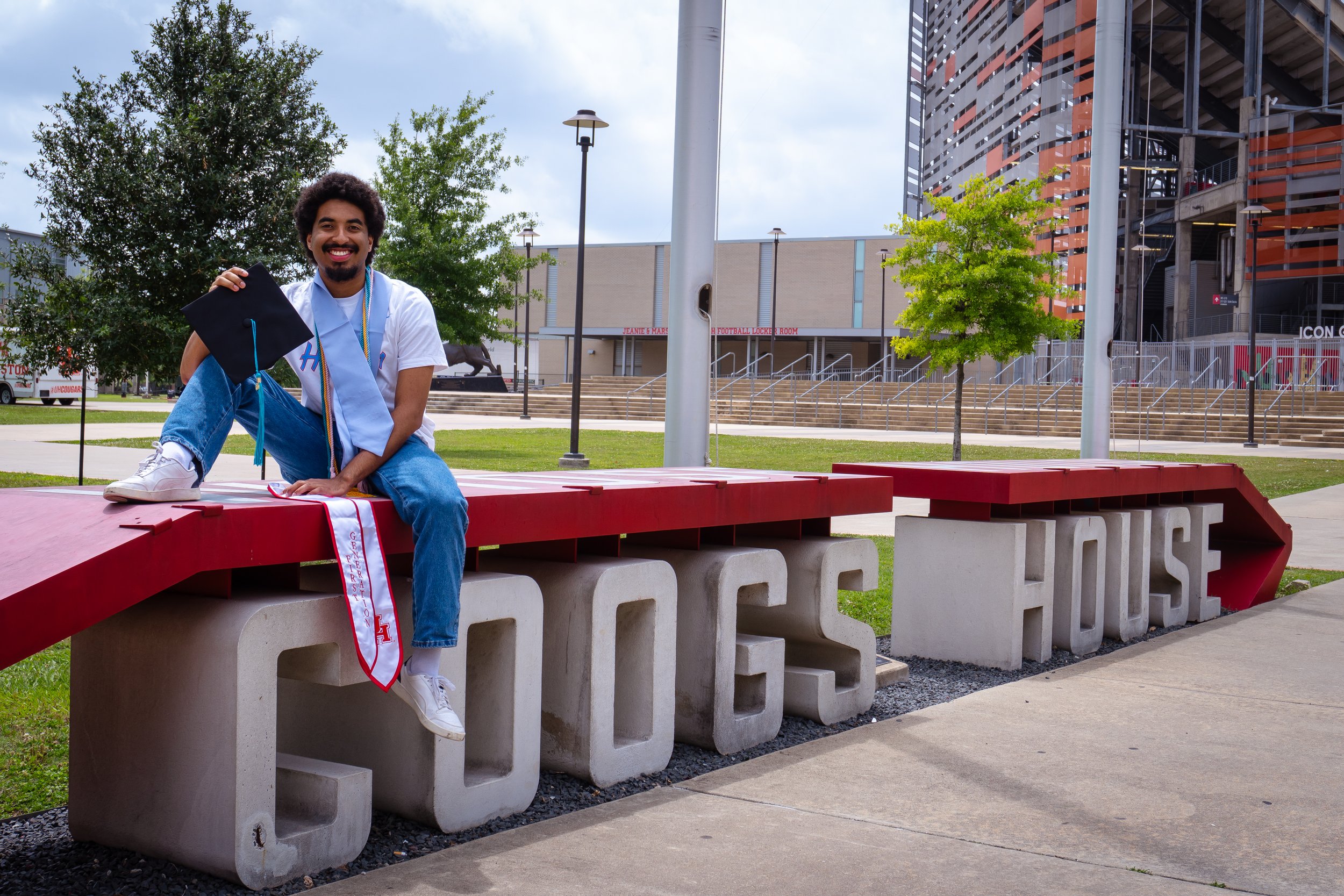 A young man sitting on a red and white university bench, holding a graduation cap, with a large building and trees in the background.