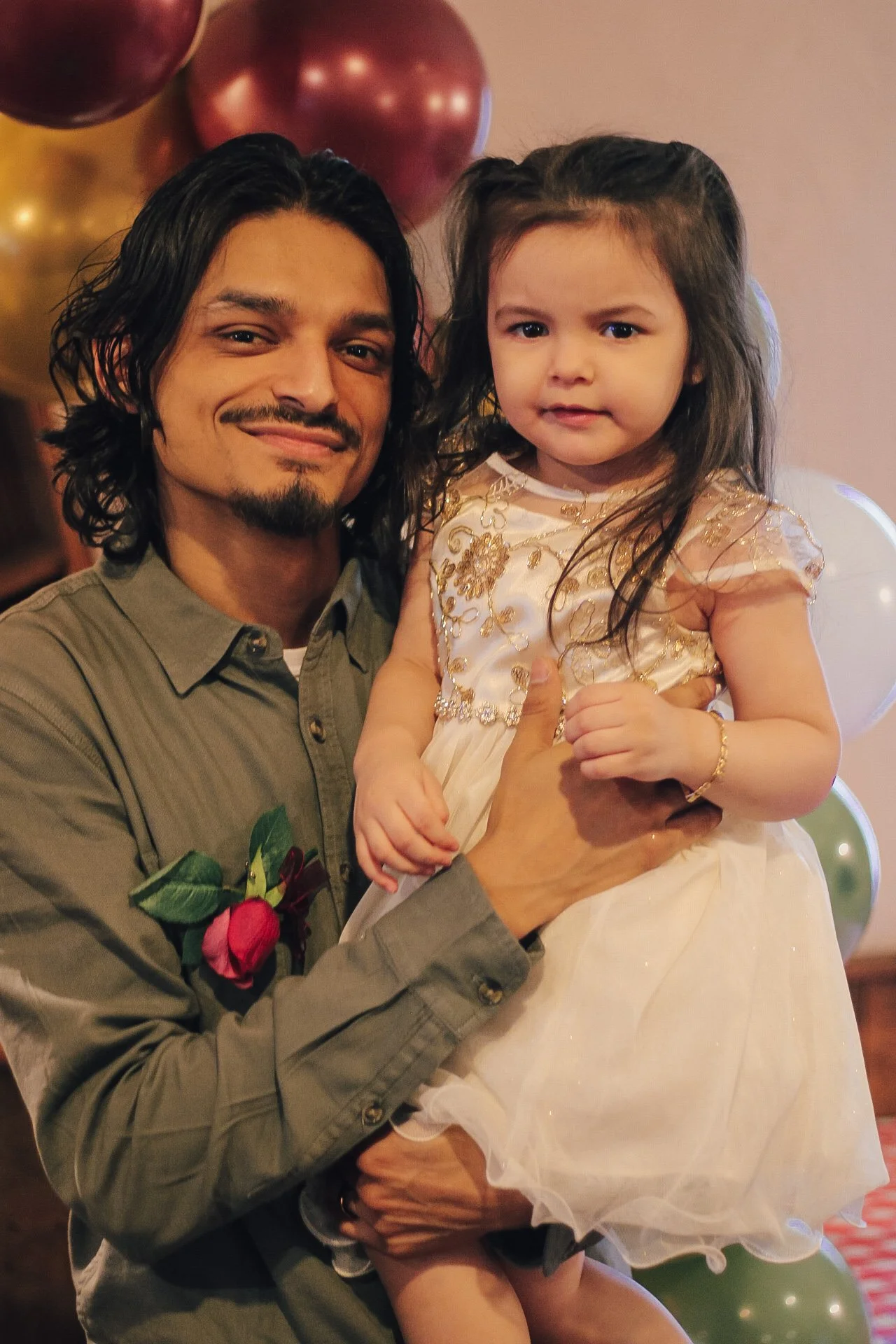 A man with shoulder-length dark hair and a goatee is holding a young girl in his arms. The girl has long dark hair and is wearing a fancy cream-colored dress with gold embroidery. They are at a celebration with balloons in the background.