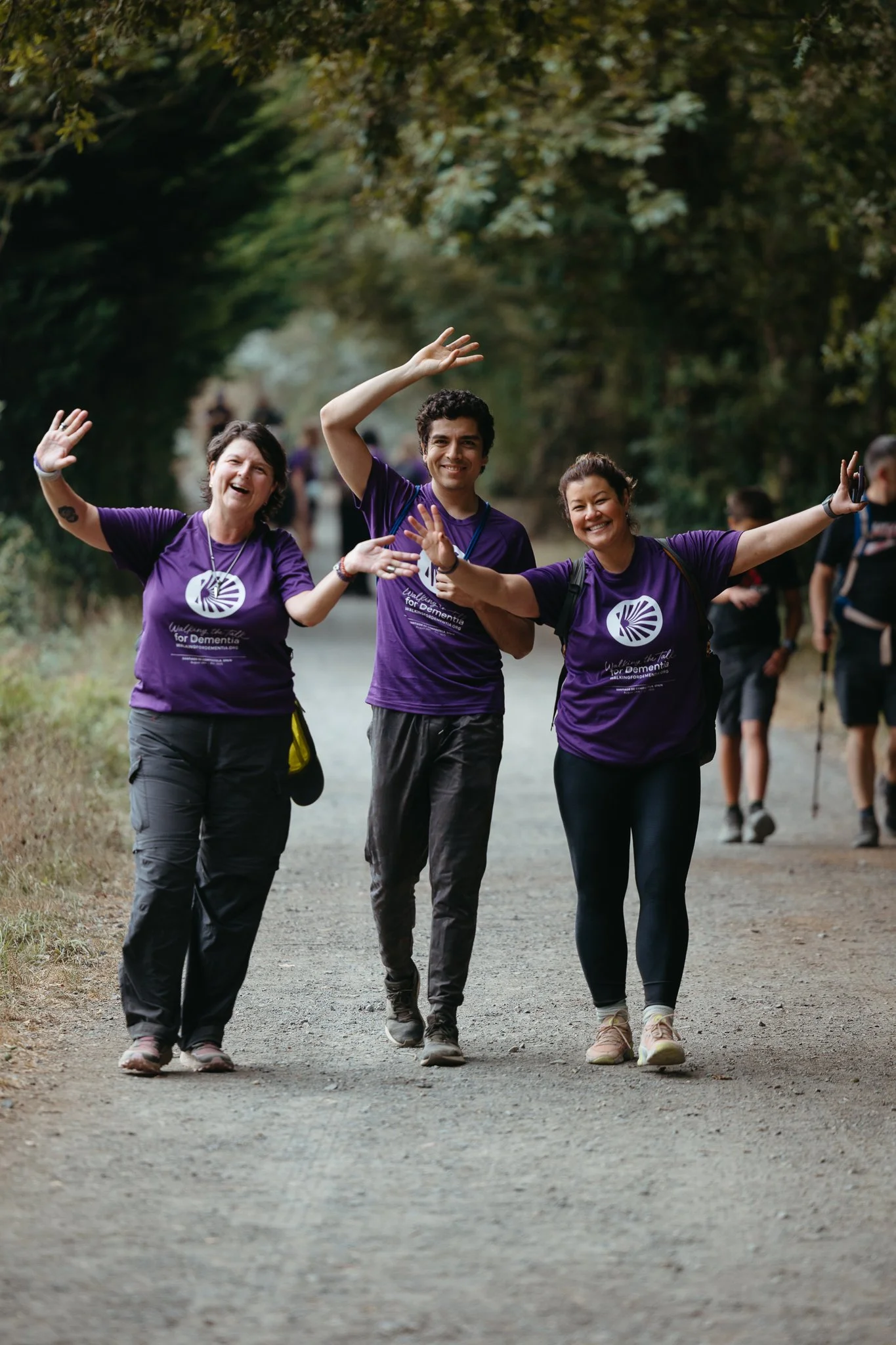 Three people smiling and waving in purple t-shirts on a dirt trail in a park.