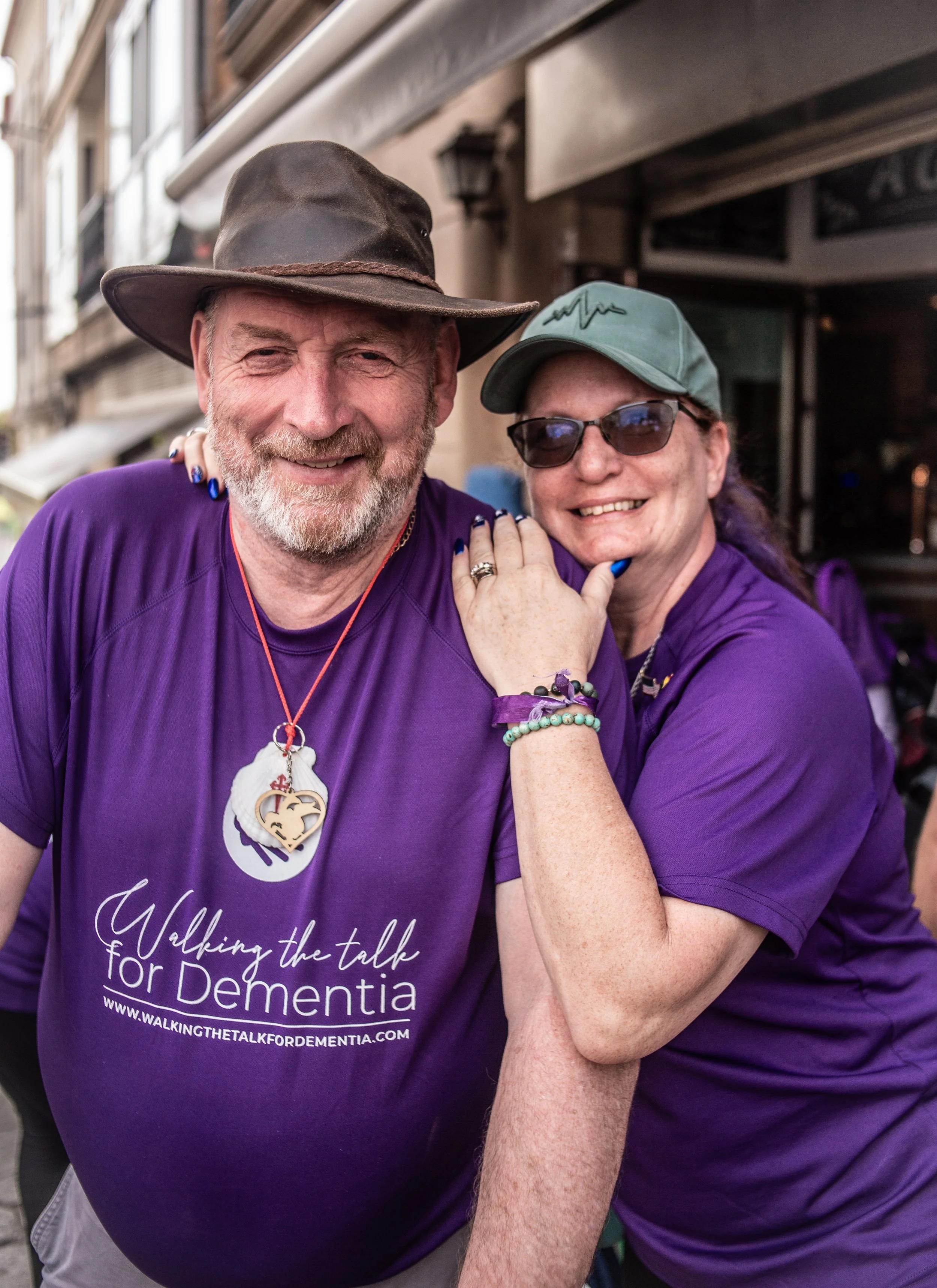 Two smiling people, a man and a woman, standing together outdoors, wearing purple t-shirts and accessories. The man's shirt supports the 'Walking the Talk for Dementia' event, and they appear happy.