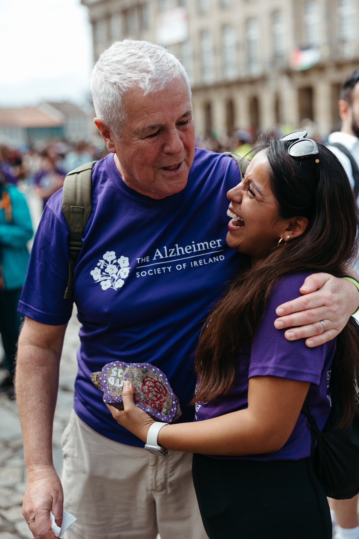 A man and woman smiling and hugging each other at an outdoor event, both wearing purple shirts related to the Alzheimer's Society of Ireland.