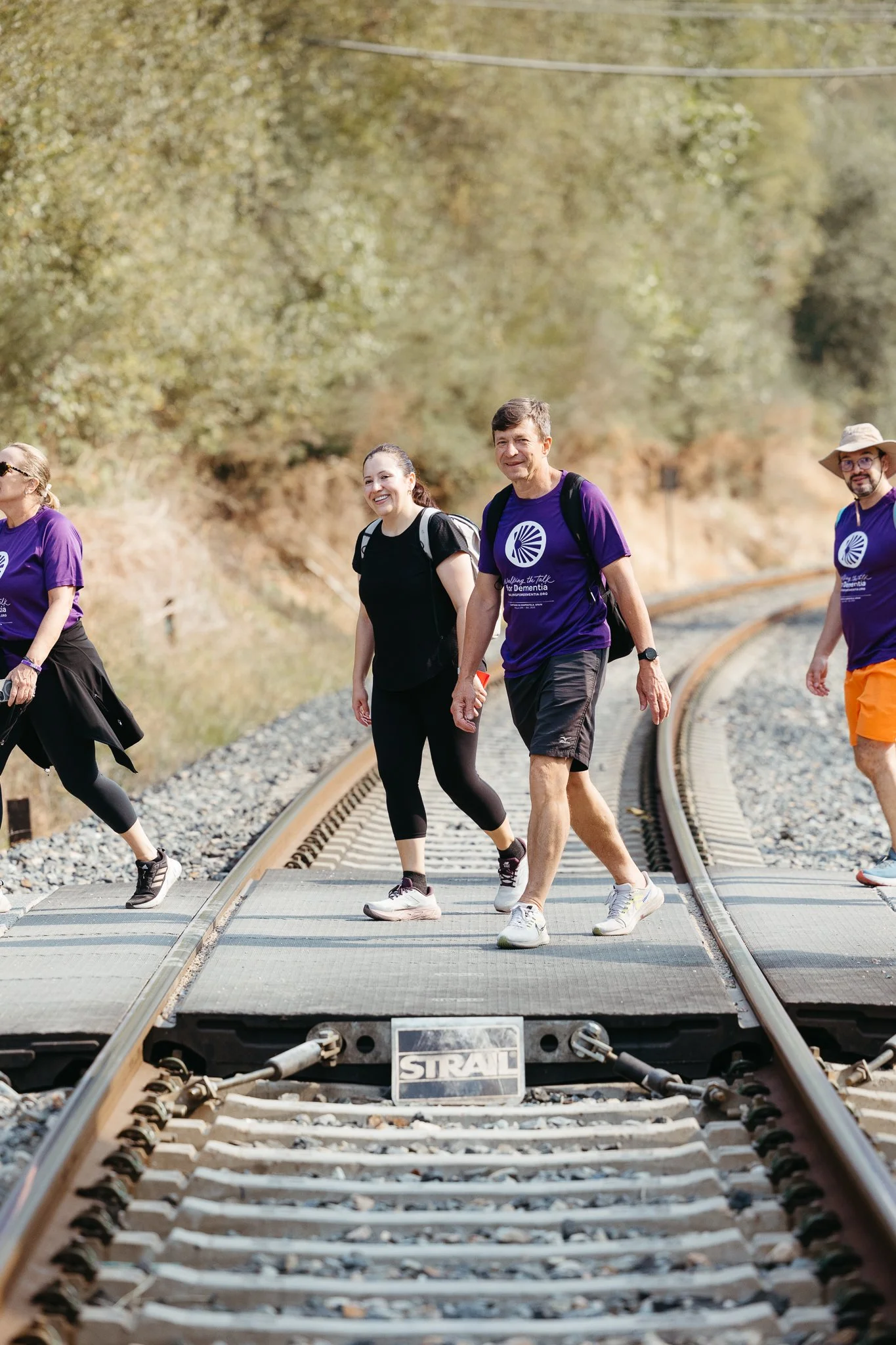 Group of people walking on a railroad track outdoors, wearing casual clothes and backpacks on a sunny day.