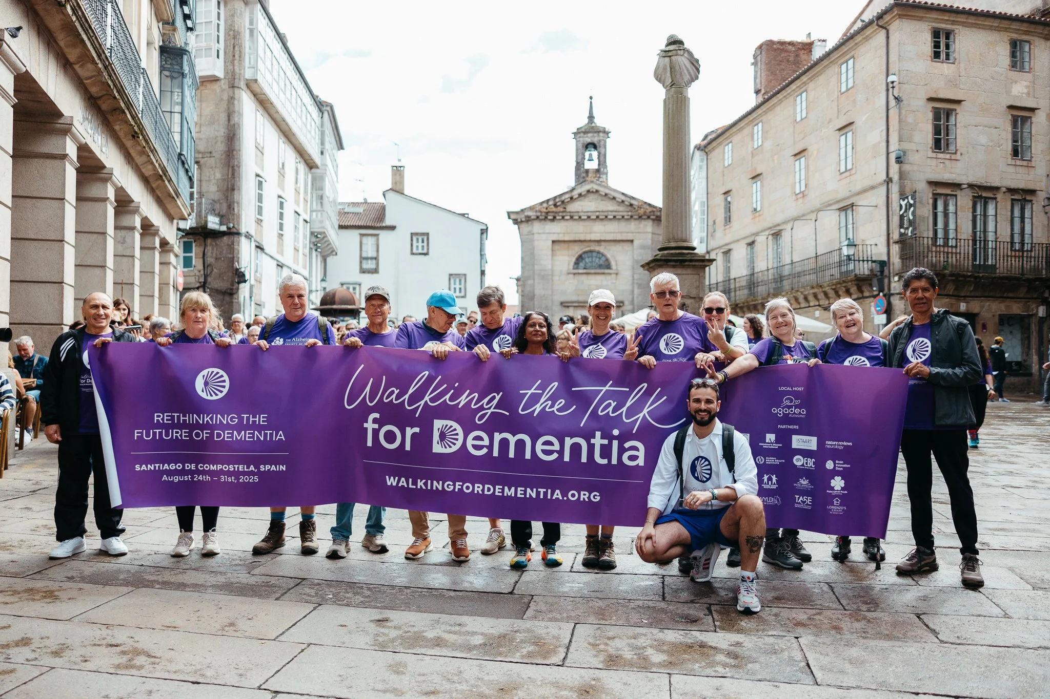 Group of people holding a purple banner that reads 'Walking the Talk for Dementia' in a historic city square. The banner also mentions 'Rethinking the Future of Dementia' in Santiago de Compostela, Spain, August 24th-31st, 2025. The group is diverse 