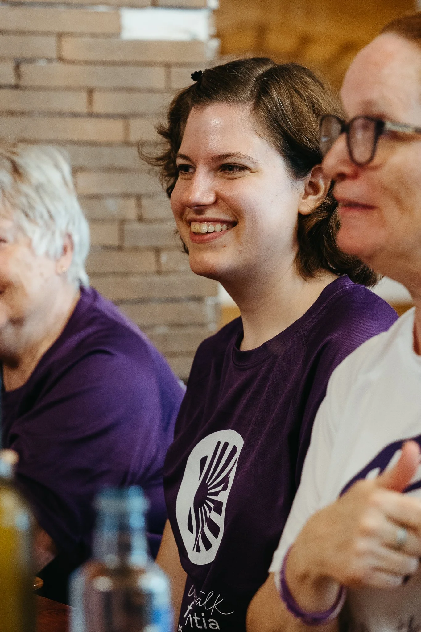 Smiling young woman with short brown hair wearing a purple T-shirt sitting at a table with elderly women, with a brick wall in the background.