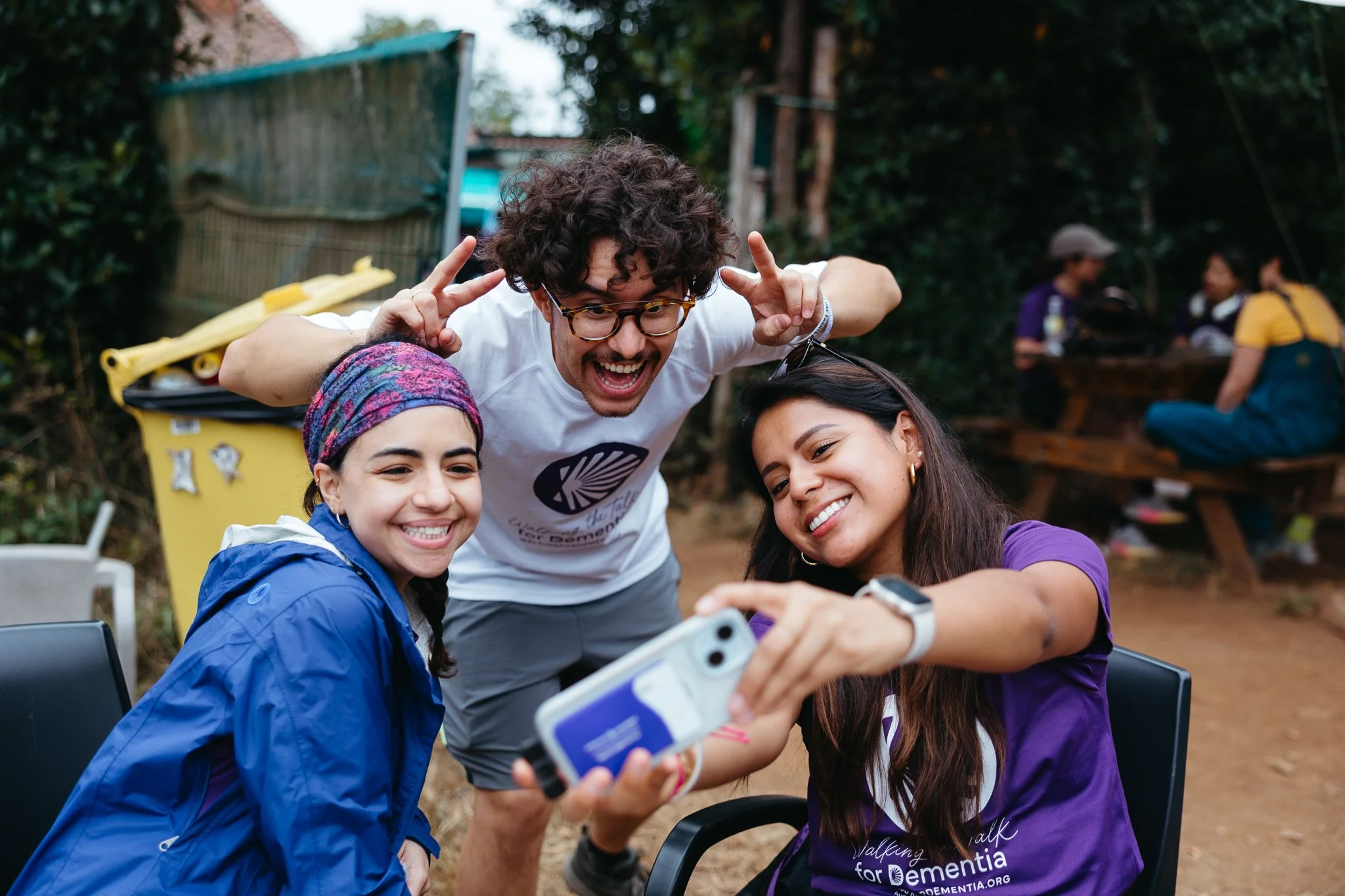 Three friends taking a selfie outdoors, smiling and making peace signs, with a group sitting at a picnic table in the background.