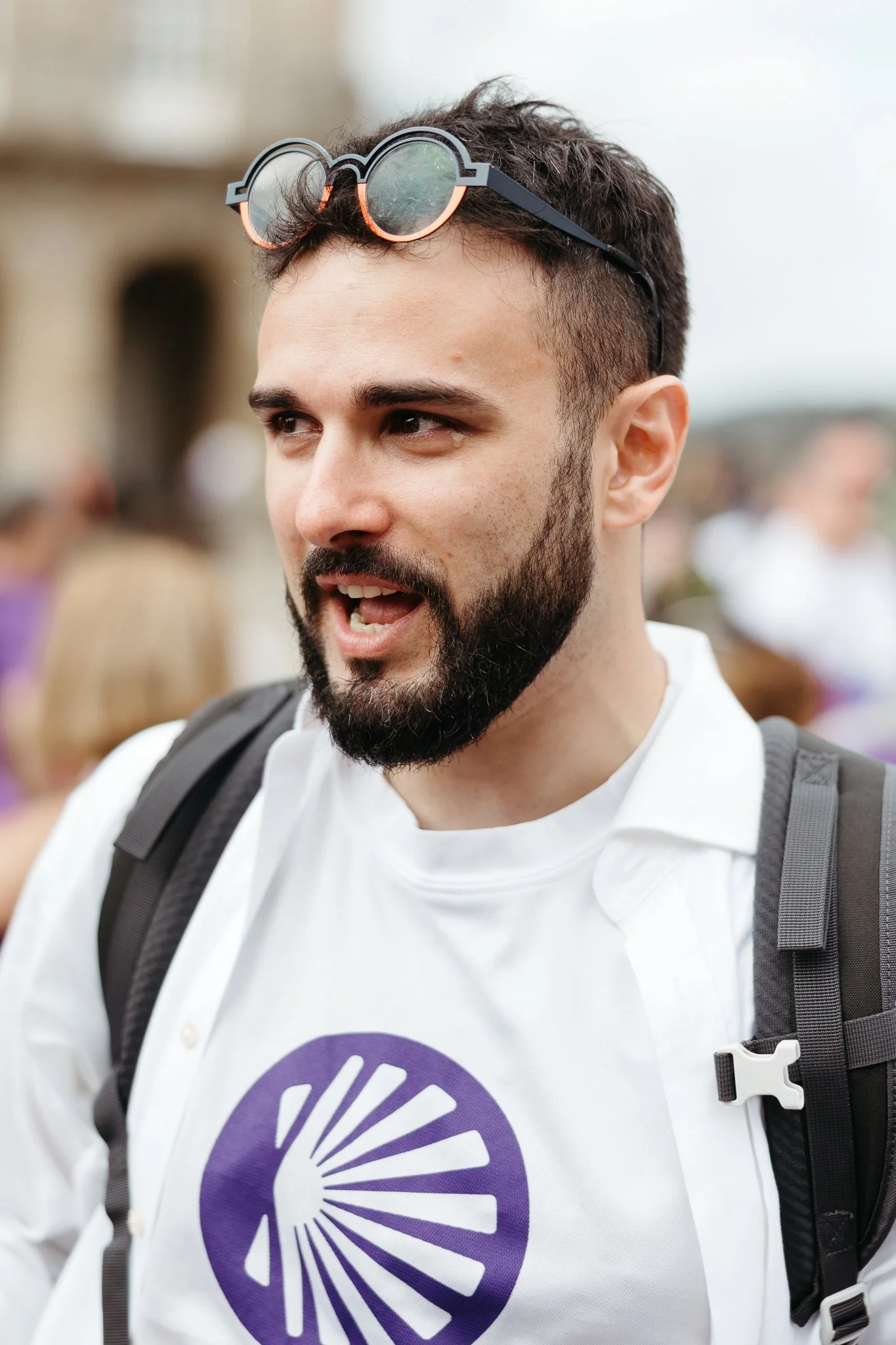 A young man with dark hair and a beard wearing sunglasses on his head, a white shirt with a purple logo, and a black backpack, outdoors.