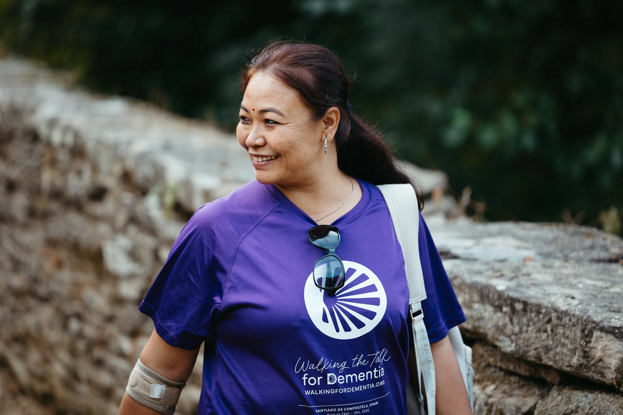 Smiling woman wearing a purple event shirt with sunglasses hanging from the neckline, walking outdoors with a stone wall and greenery in the background.
