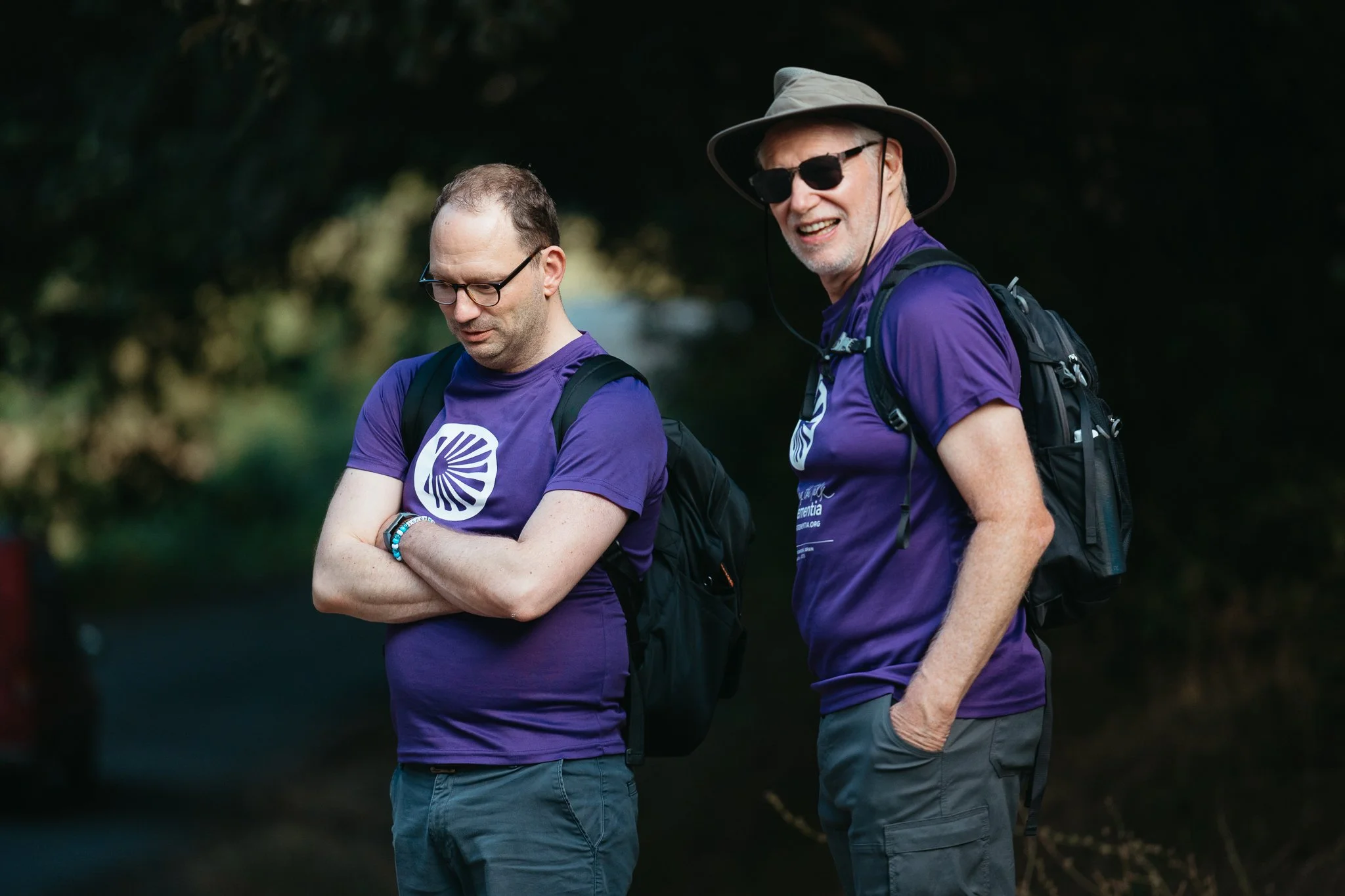 Two men wearing purple shirts and backpacks standing outdoors in a wooded area, one with arms crossed and looking down, the other smiling wearing sunglasses and a wide-brimmed hat.