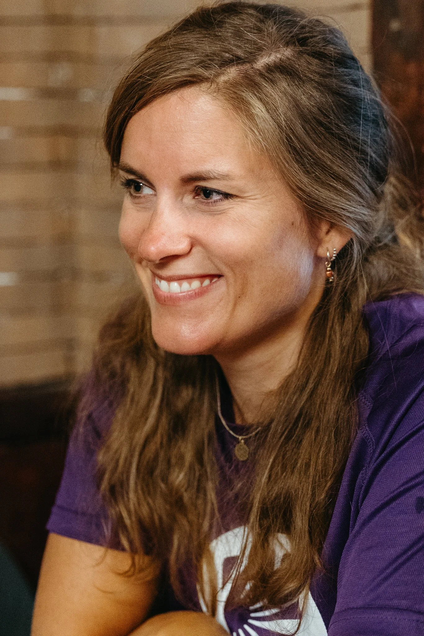 Close-up of a smiling woman with long brown hair, wearing earrings, a necklace, and a purple shirt.