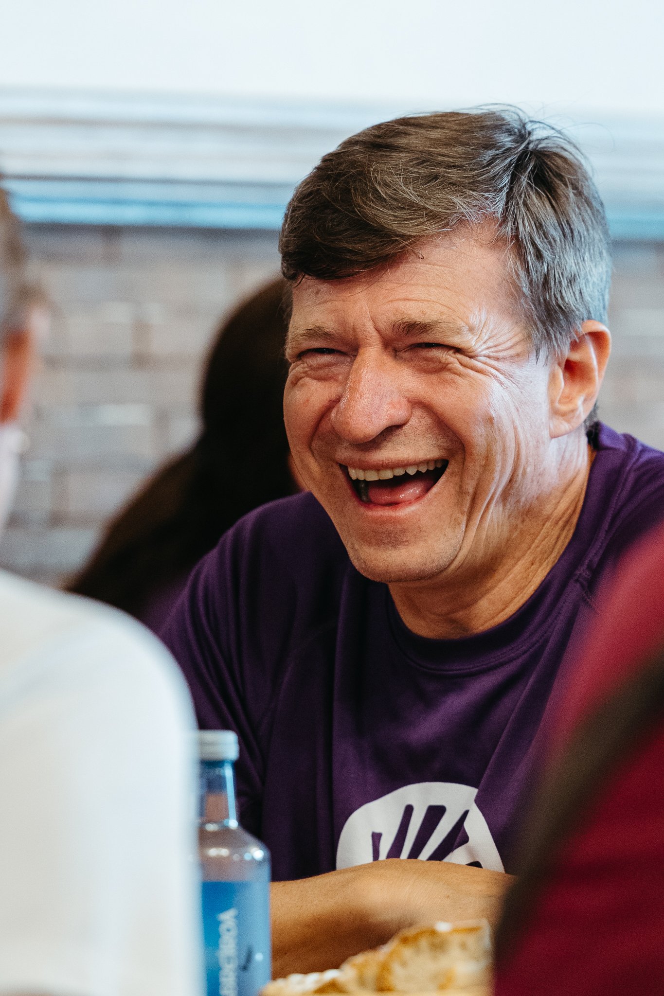 A middle-aged man with gray hair laughing. He is wearing a purple shirt and is sitting at a table with a bottle of water and some food.