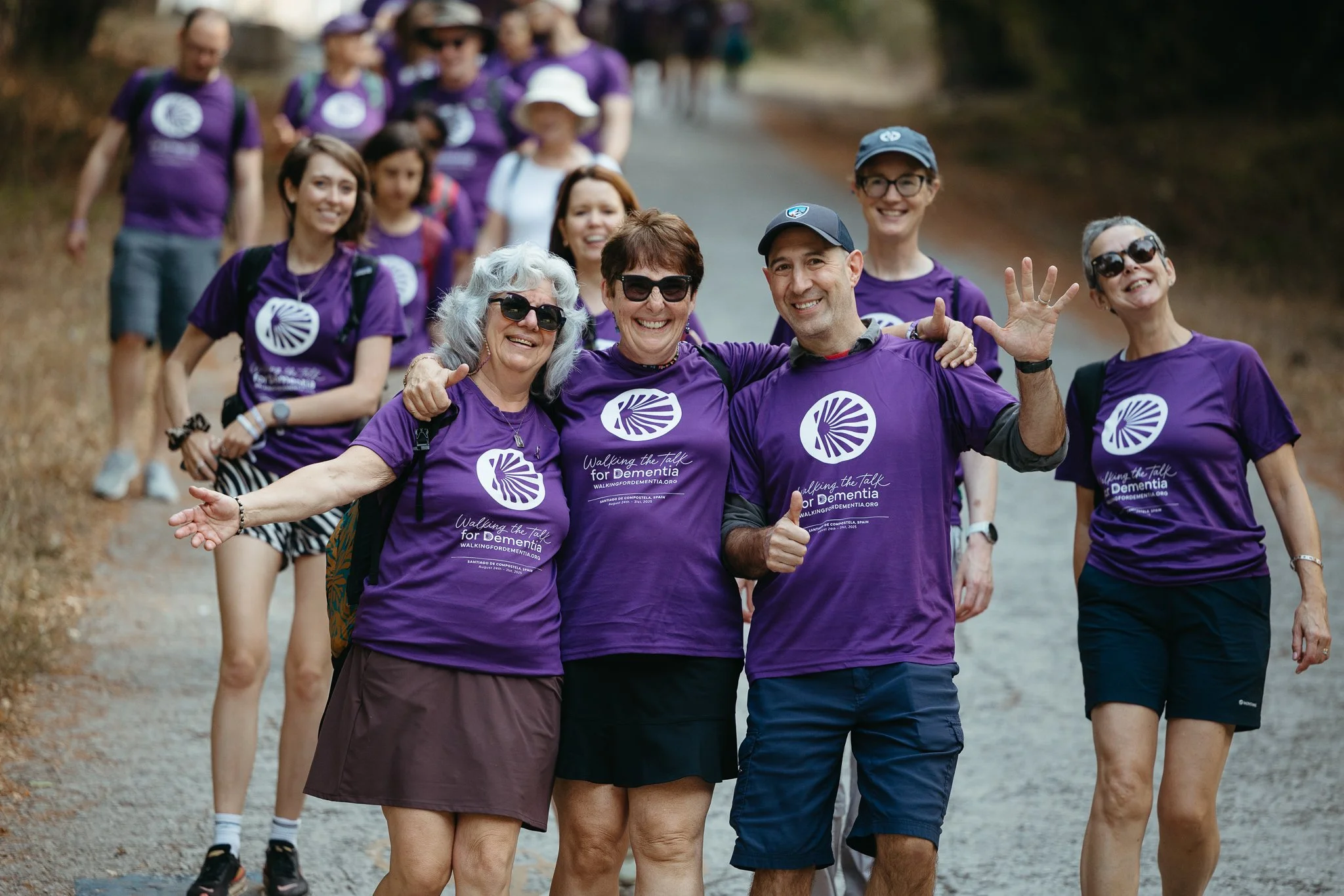 Group of people walking outdoors, wearing matching purple shirts supporting Dementia awareness, walking on a gravel path surrounded by nature.