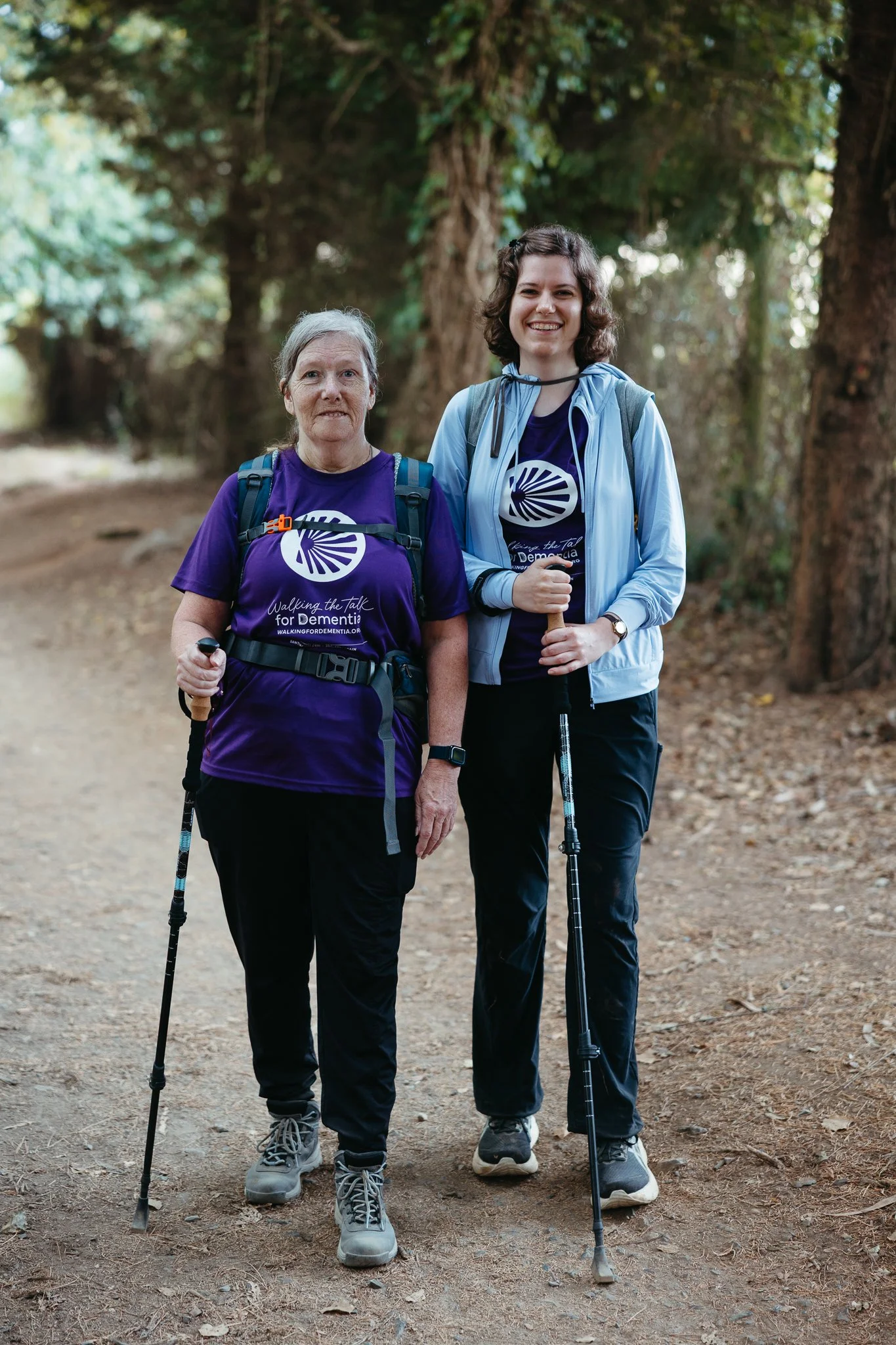 Two women hiking on a dirt path through a forest, wearing purple shirts with a logo and holding trekking poles, smiling at the camera.