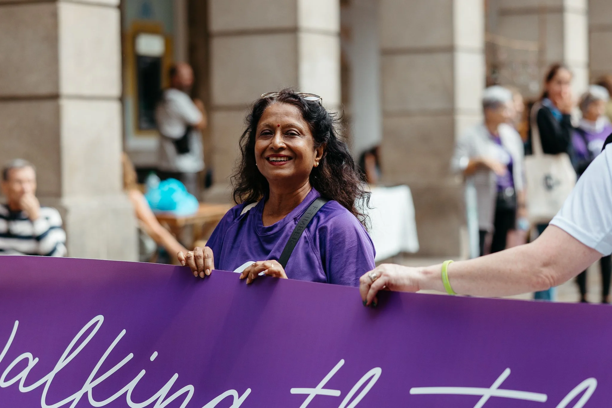 A woman with dark curly hair and a purple shirt, smiling and holding a purple banner at a public event, with people in the background.