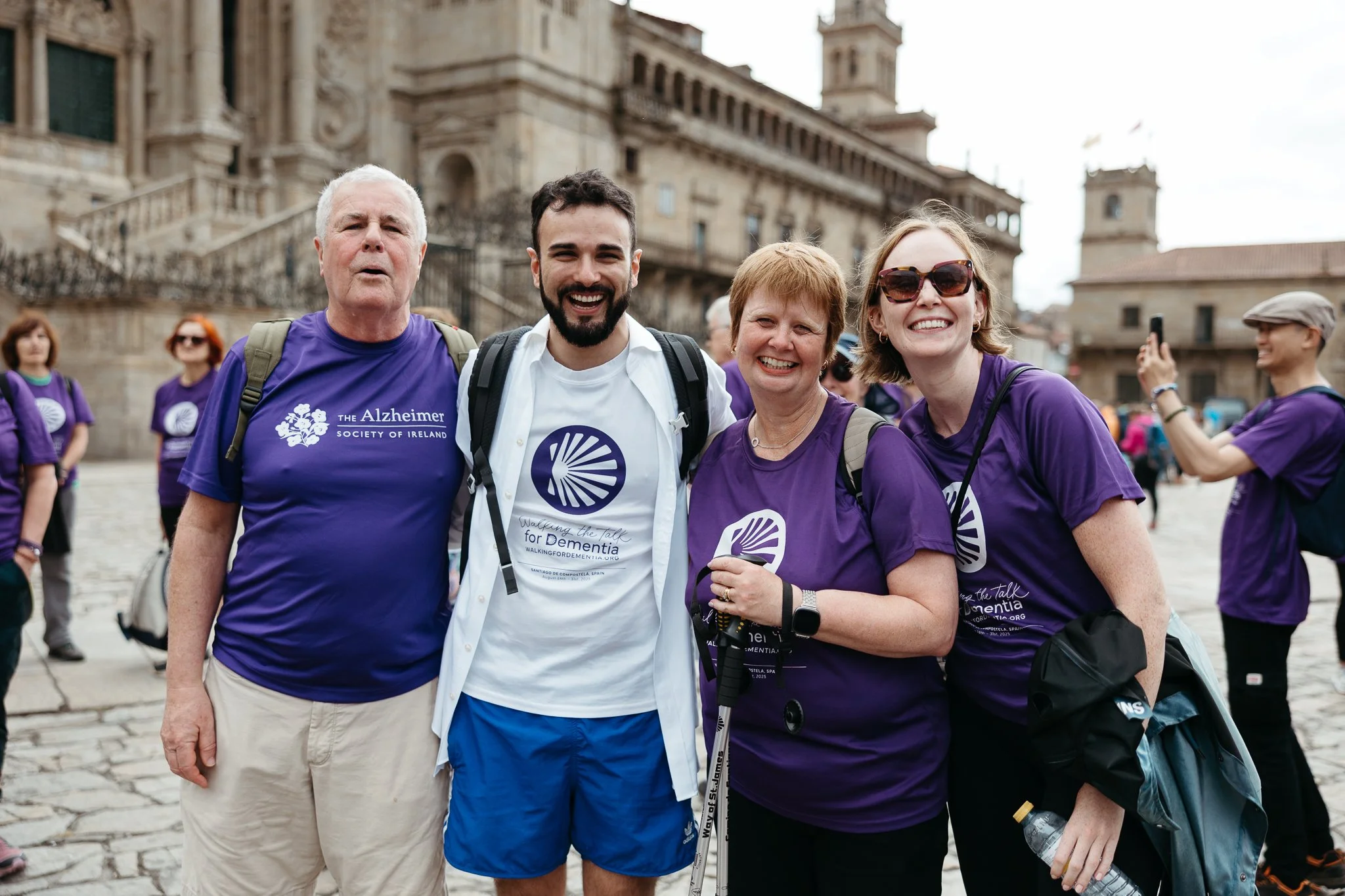 Group of four smiling people standing together outdoors, some wearing purple Alzheimer Society shirts, with a historic building in the background, during a dementia awareness walk event.