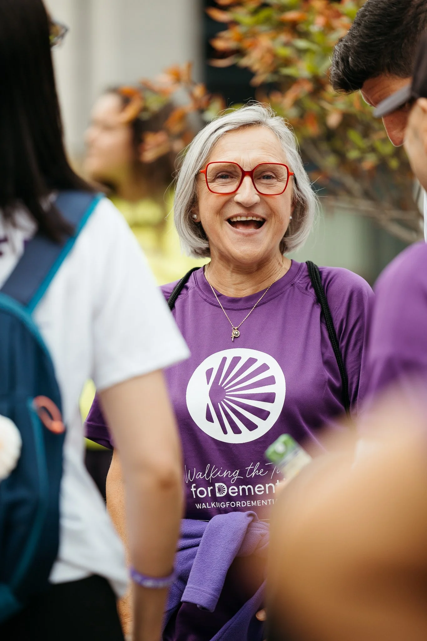 An elderly woman with gray hair and red glasses smiling and talking to a group at an outdoor event. She is wearing a purple shirt with a logo and text for 'Walking the Talk for Dementia'.