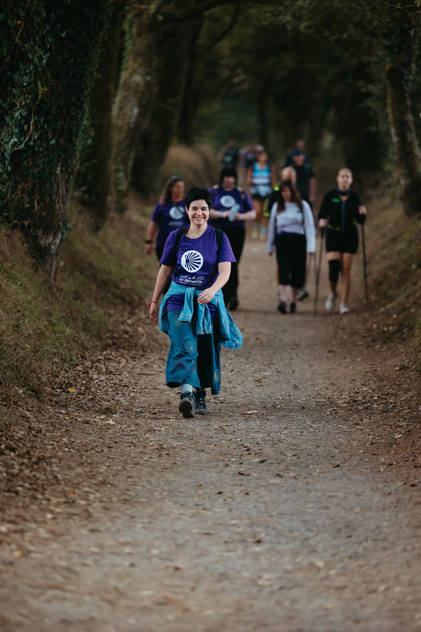 Group of people walking along a dirt trail in a forested area, with one woman smiling at the camera in the foreground.