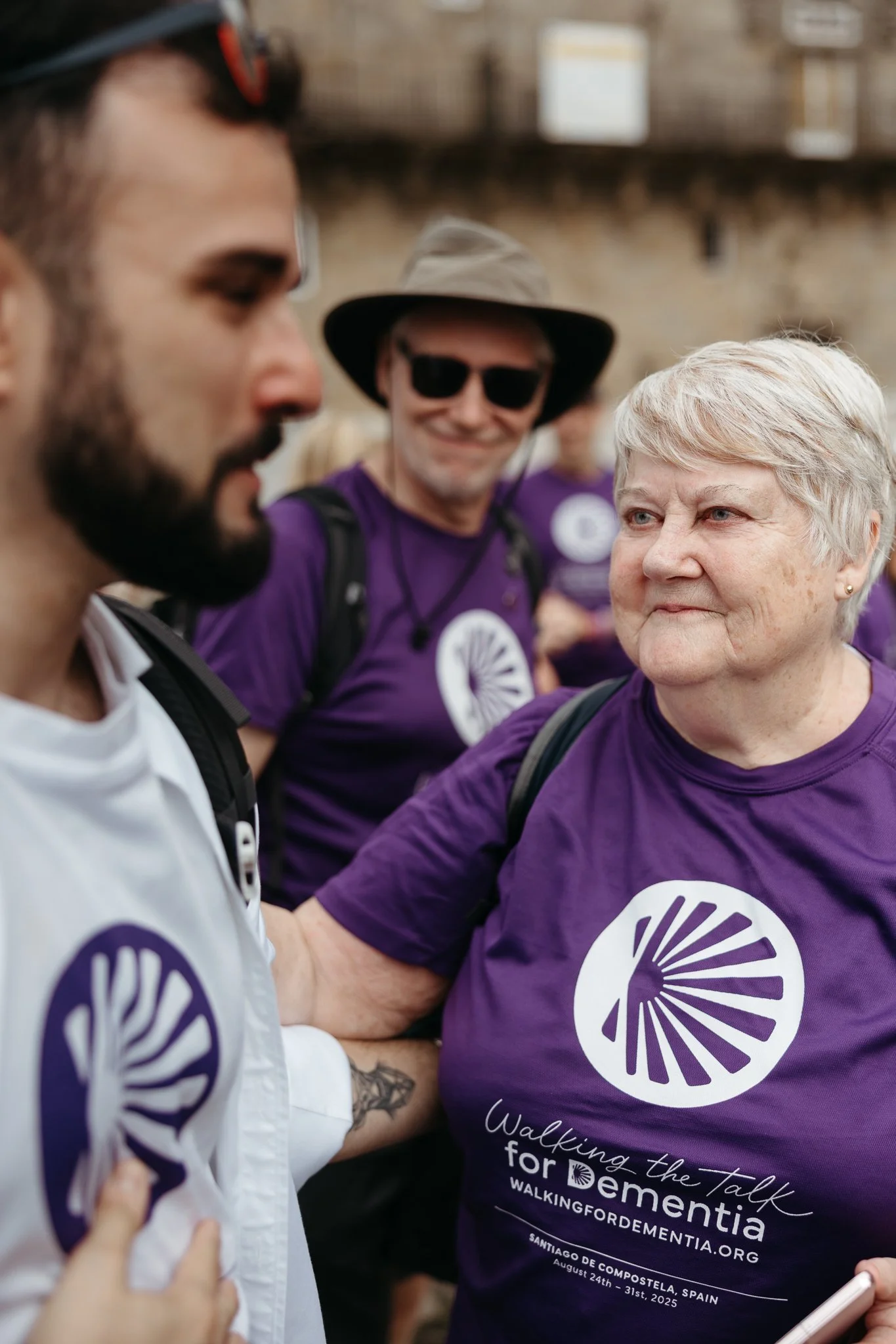 A woman wearing a purple T-shirt with a logo and text raising her hand to greet a man, with a background of people wearing similar purple shirts and a man in sunglasses and a hat smiling.