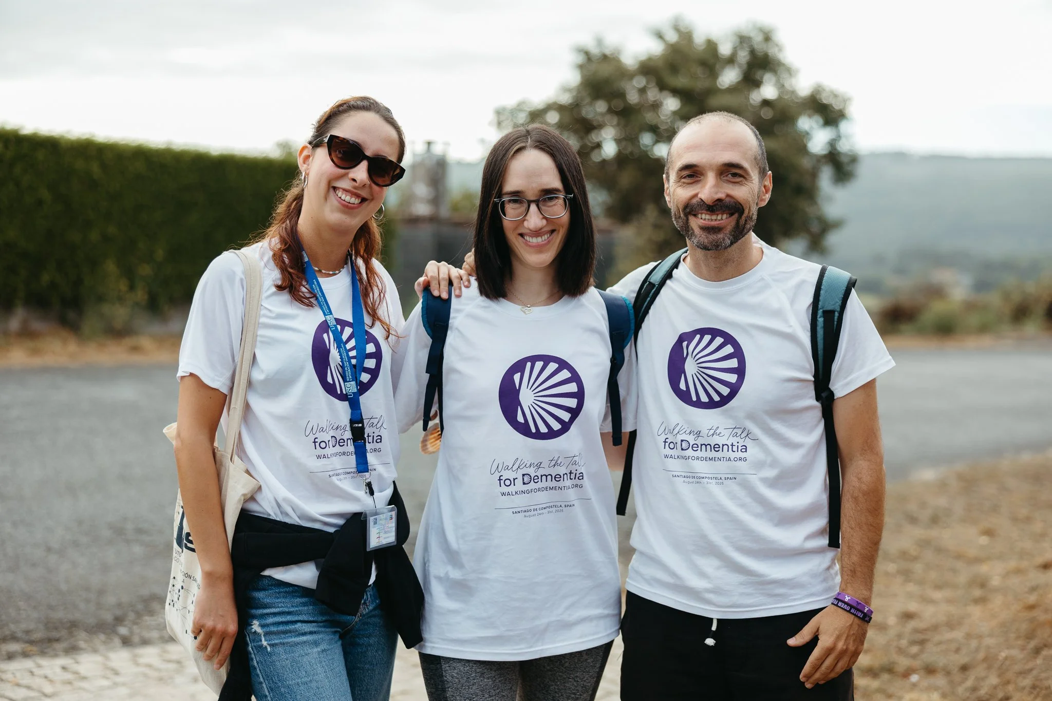 Three smiling people wearing white T-shirts with a purple circular logo, standing outdoors with a blurred background of trees and sky.