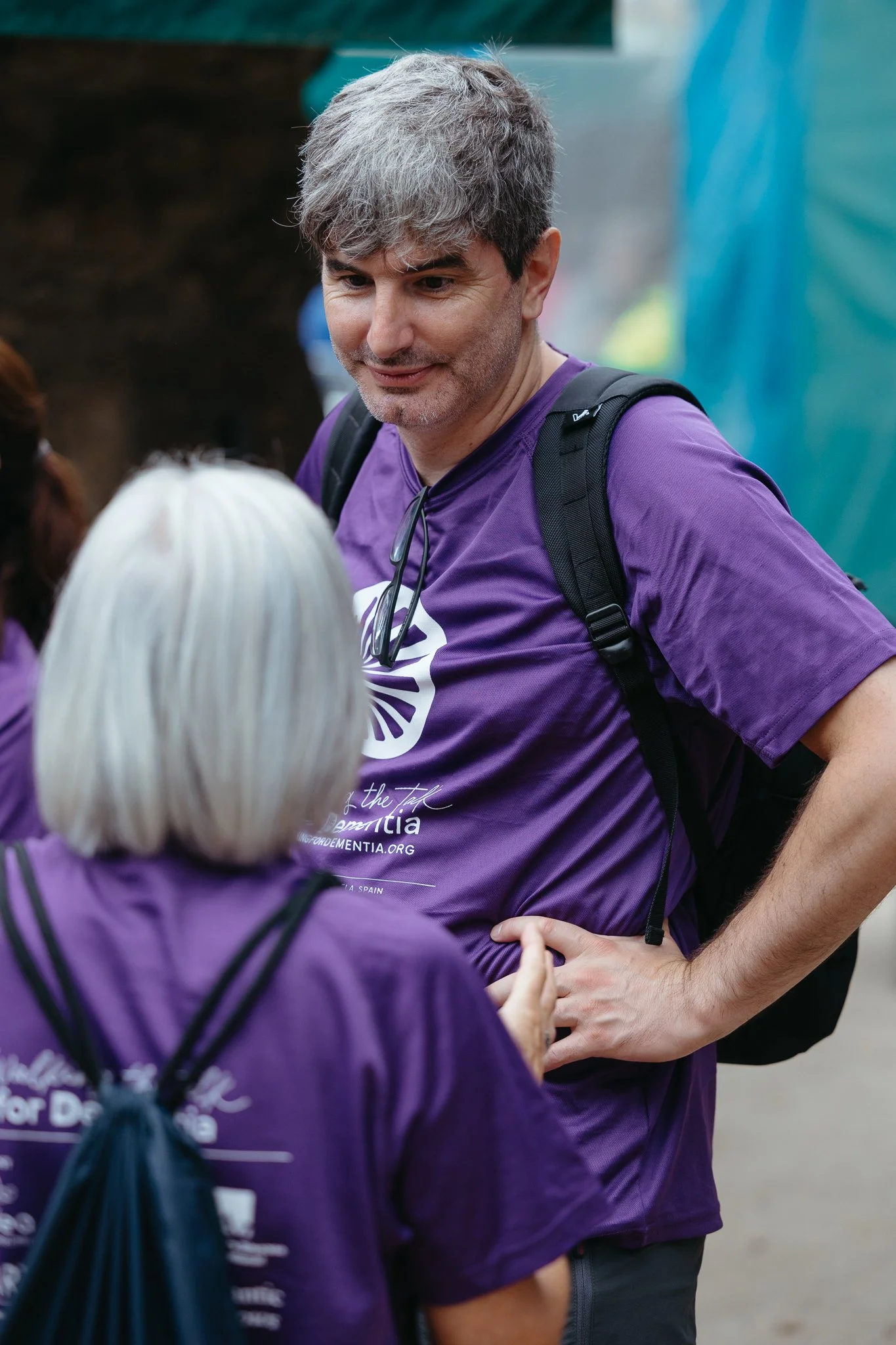 A man with gray hair and a beard, wearing a purple T-shirt and a backpack, engaged in conversation with a woman with white hair, also wearing a purple T-shirt. They are outside, connected to a comfort or charity event.