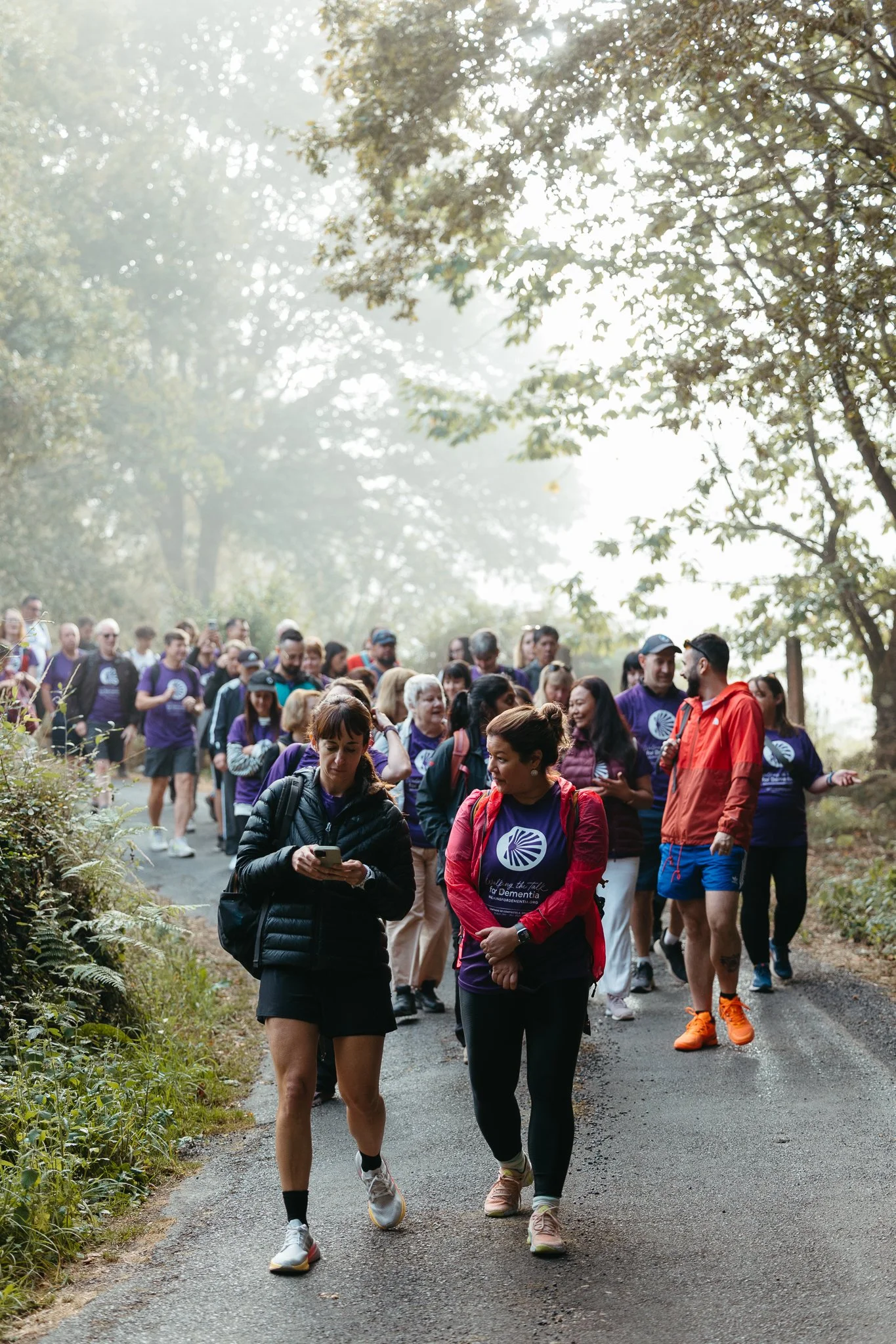 A large group of people walking on a forest trail during daytime, some wearing purple shirts with a logo, others in various athletic clothing, surrounded by tall trees and sunlight filtering through the leaves.