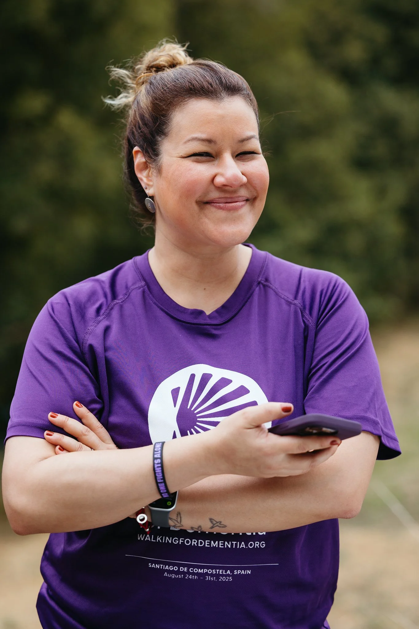A woman smiling outdoors, wearing a purple t-shirt with event information, holding a smartphone, with arms crossed, in front of a blurred green background.