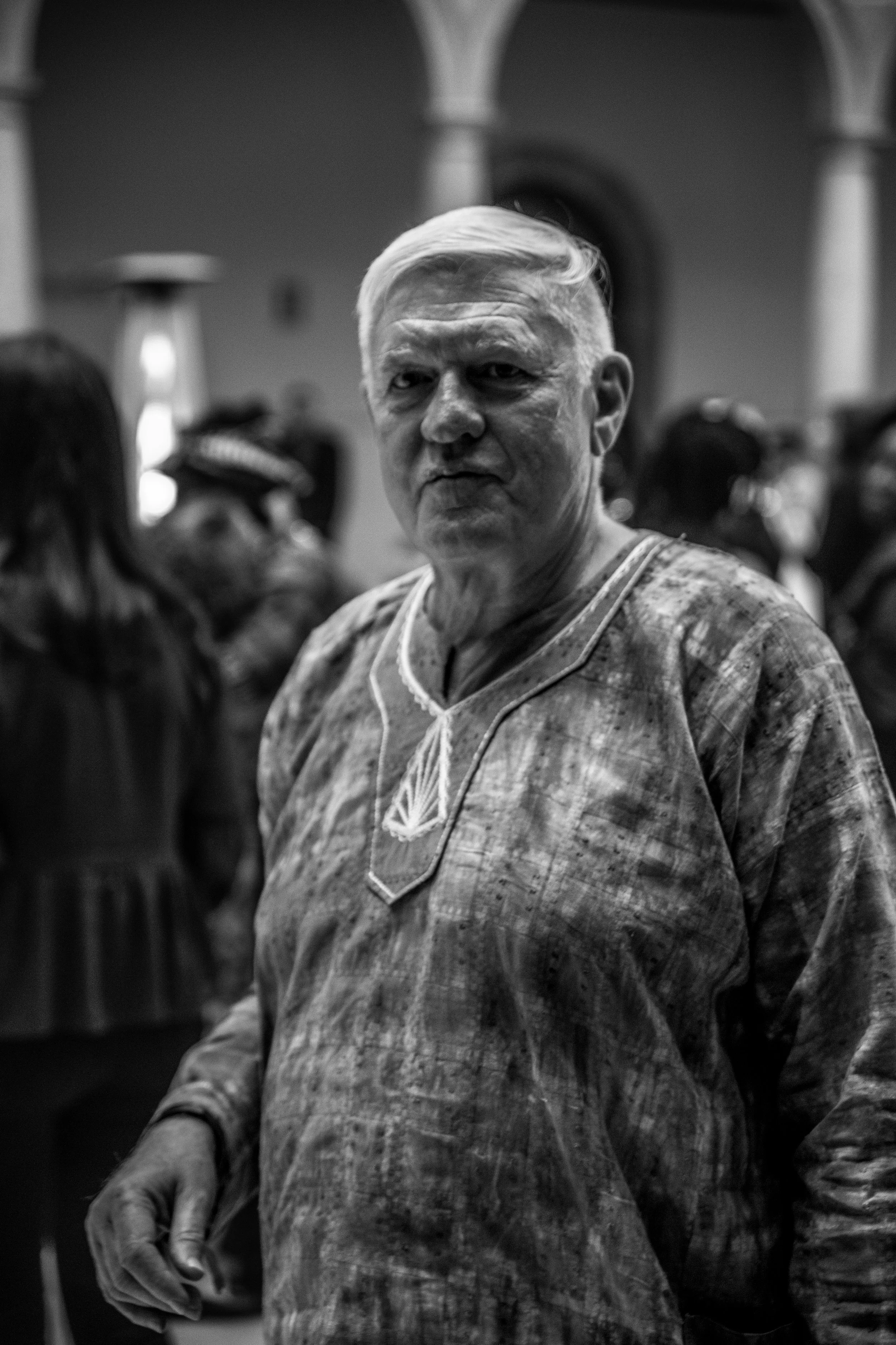 Black and white photo of an elderly man with short white hair wearing a patterned traditional African shirt, standing among a crowd indoors.