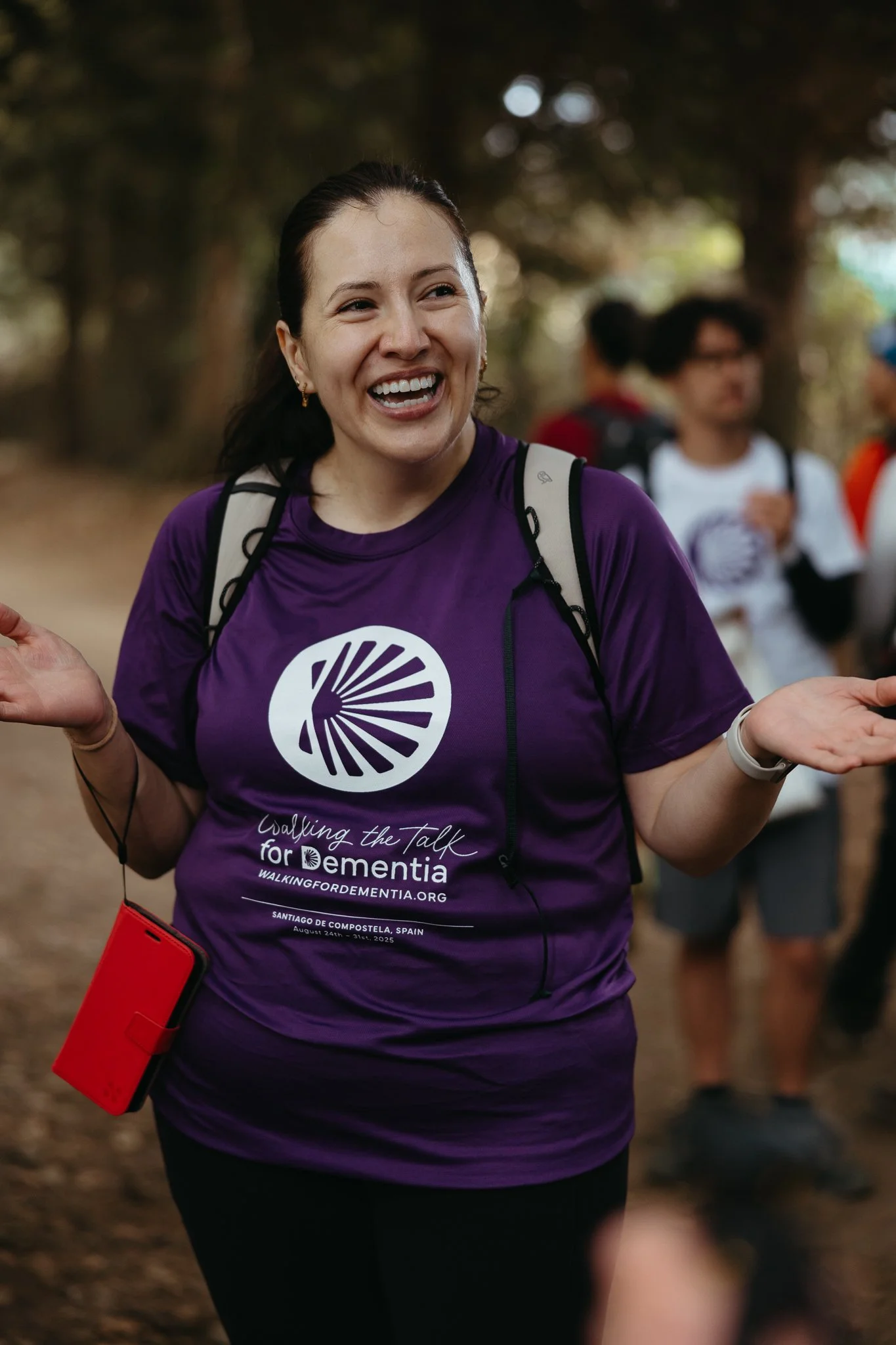 A woman smiling and gesturing with her hands while participating in a walk or event in a wooded area. She is wearing a purple event shirt that promotes walking for dementia, with several other people in the background.