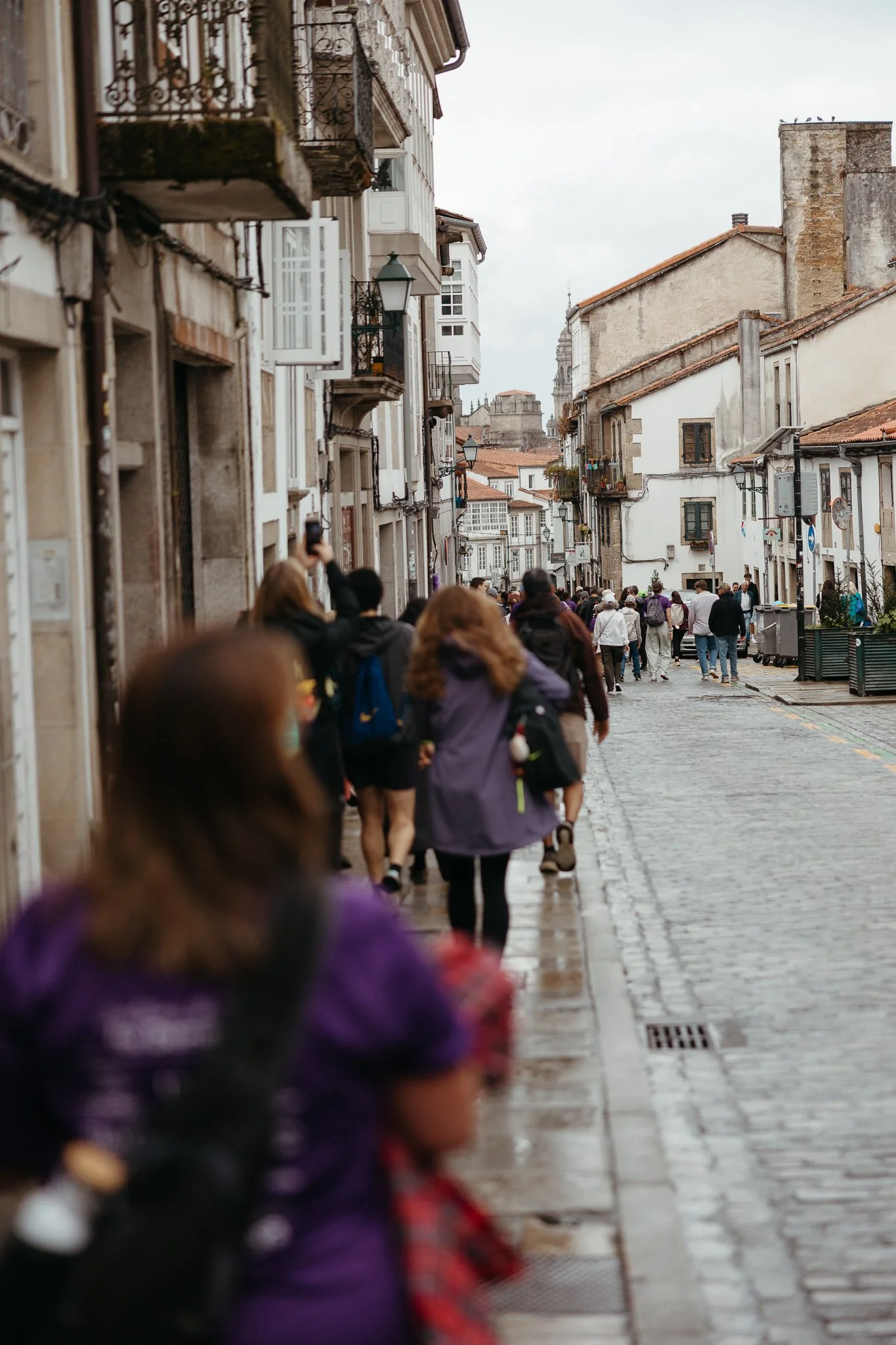 A rainy street scene in a European city with pedestrians walking along a cobblestone road, surrounded by historic buildings with balconies and street lamps, under a cloudy sky.