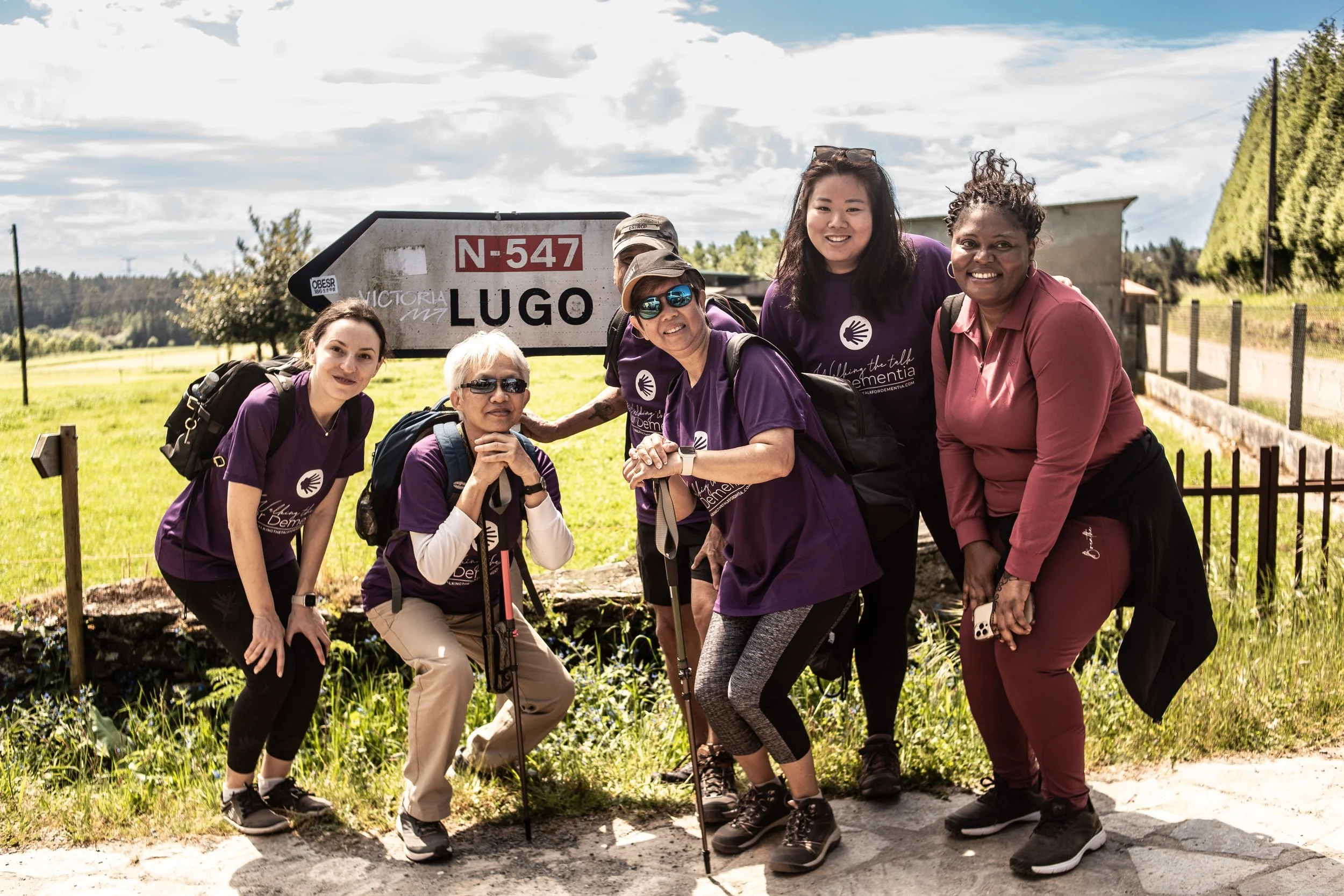 Group of six diverse women posing outdoors in front of a road sign that reads 'N-547 Lugo' in a rural area with greenery and a cloudy sky.