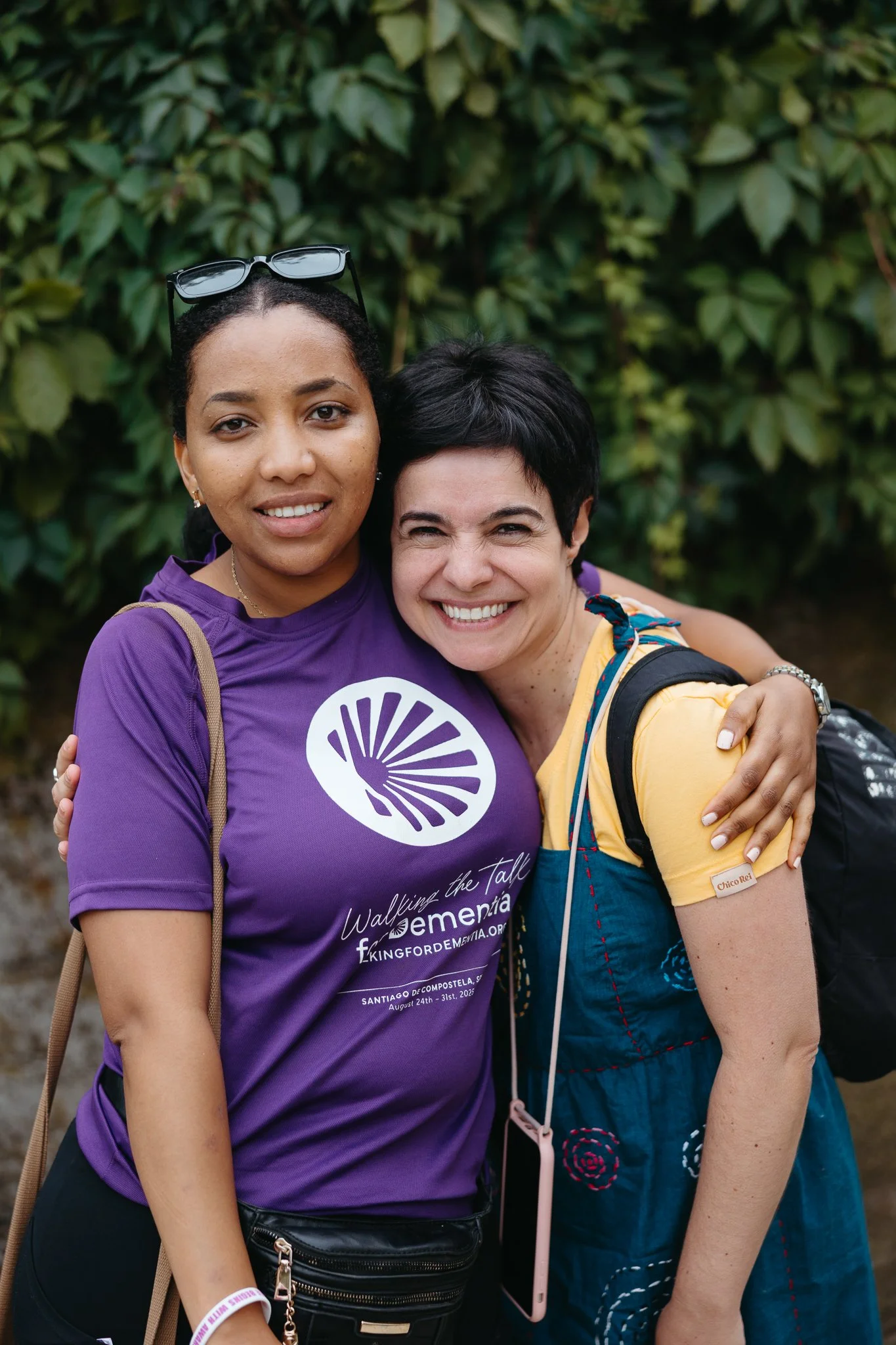 Two women hugging outdoors, smiling, with green foliage in the background. One woman wears a purple shirt with a logo and text, and the other woman wears a yellow shirt and blue overall.