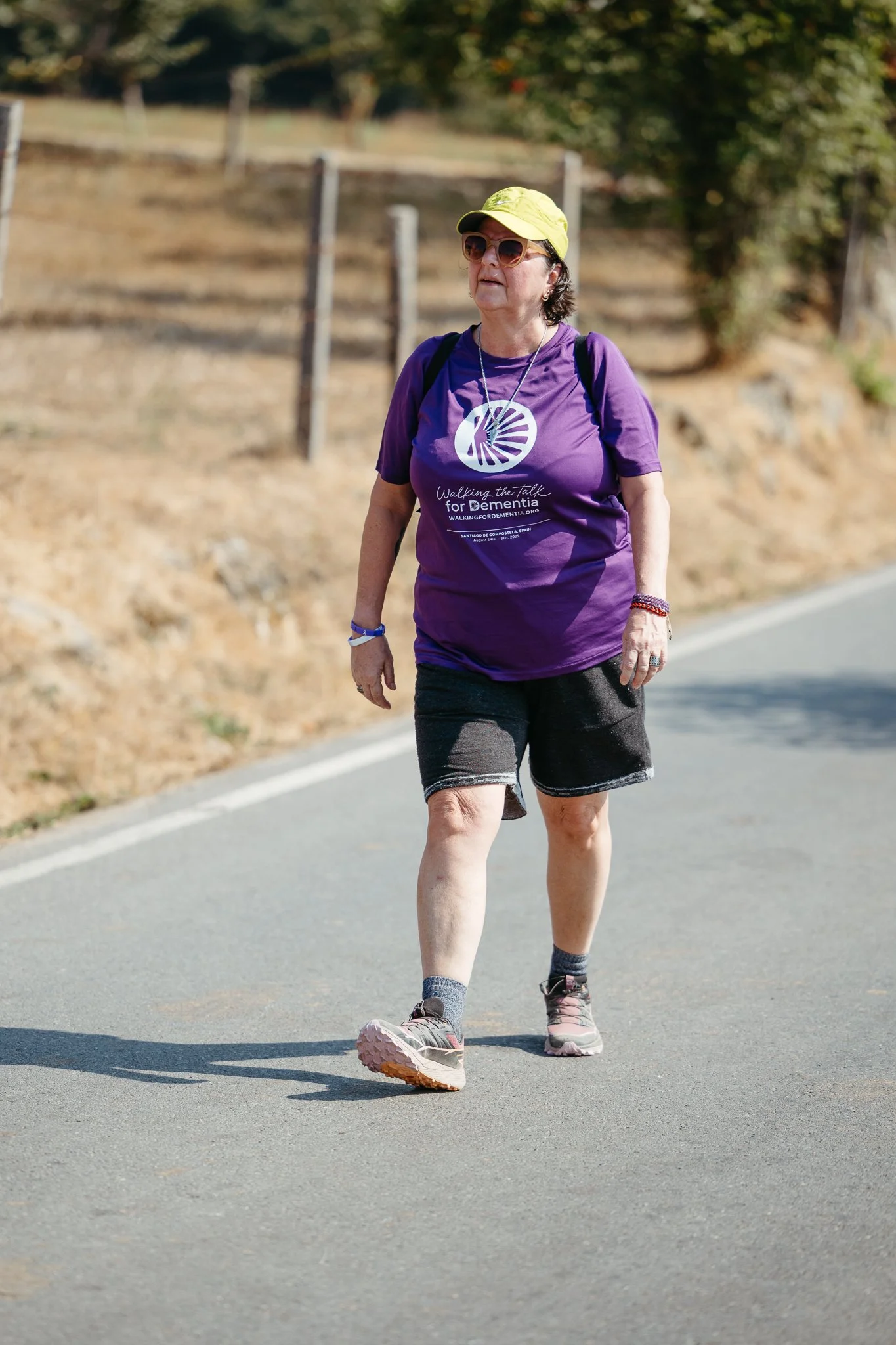 A woman walking outdoors on a paved path during daytime, wearing a purple t-shirt, black shorts, gray and pink hiking shoes, sunglasses, and a yellow cap.