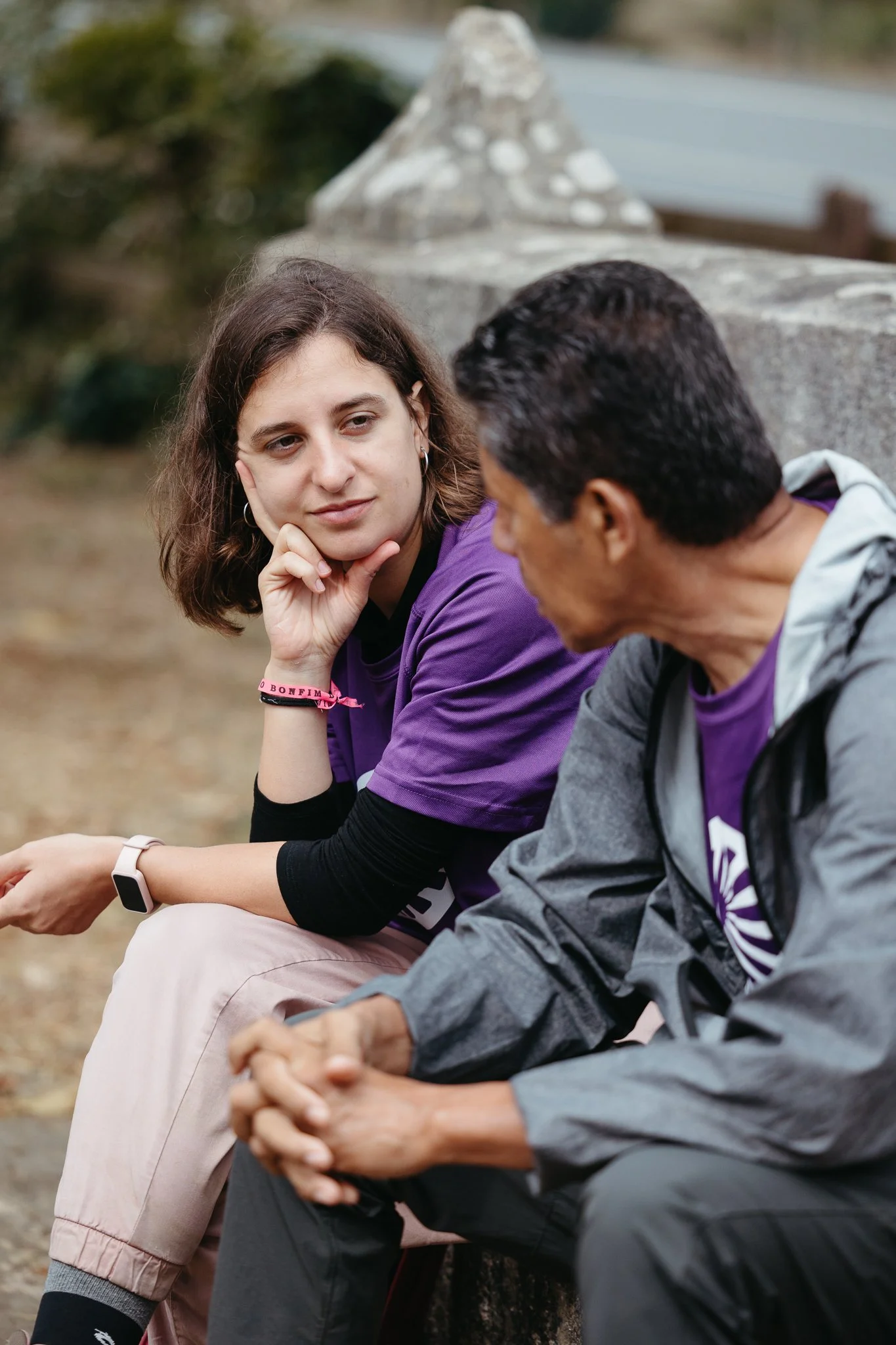 A young woman with shoulder-length brown hair and a man with short dark hair sitting outdoors, engaged in conversation.