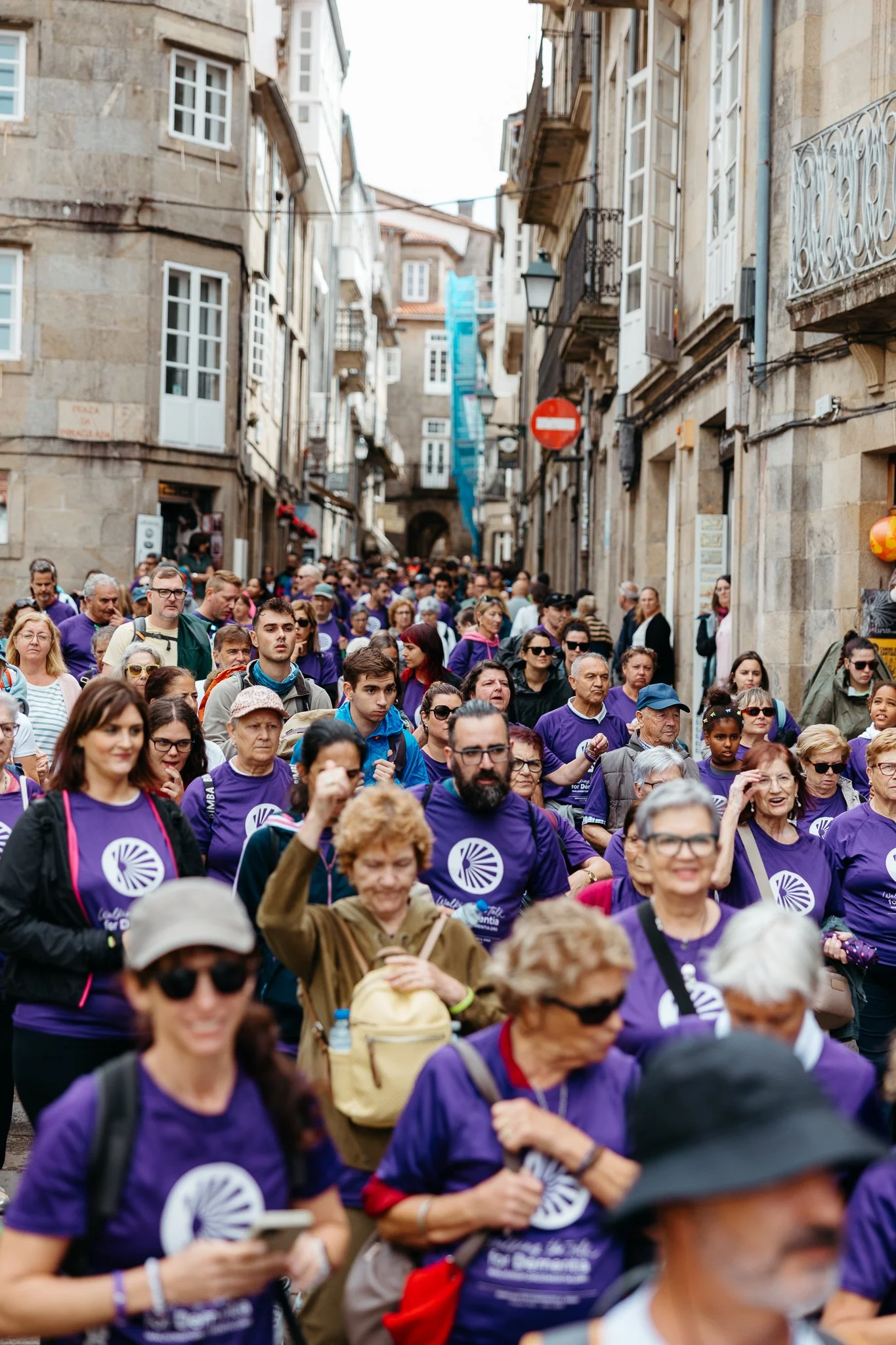 Crowd of people participating in a street event or protest, many wearing purple shirts, walking down a narrow city street.
