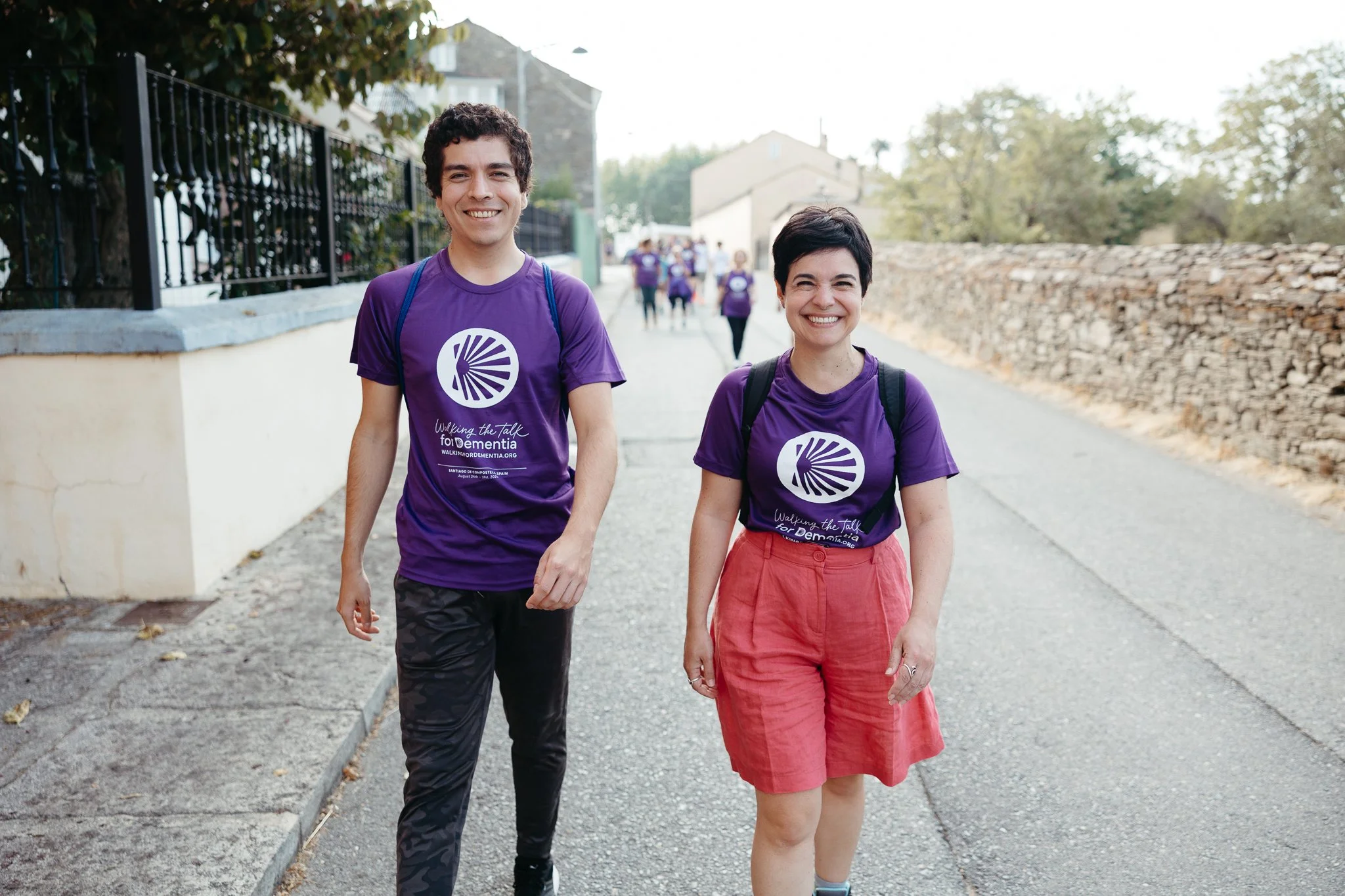 Two smiling people walking outdoors during the daytime, wearing matching purple shirts with a logo and text supporting dementia awareness, with other people in similar shirts in the background.