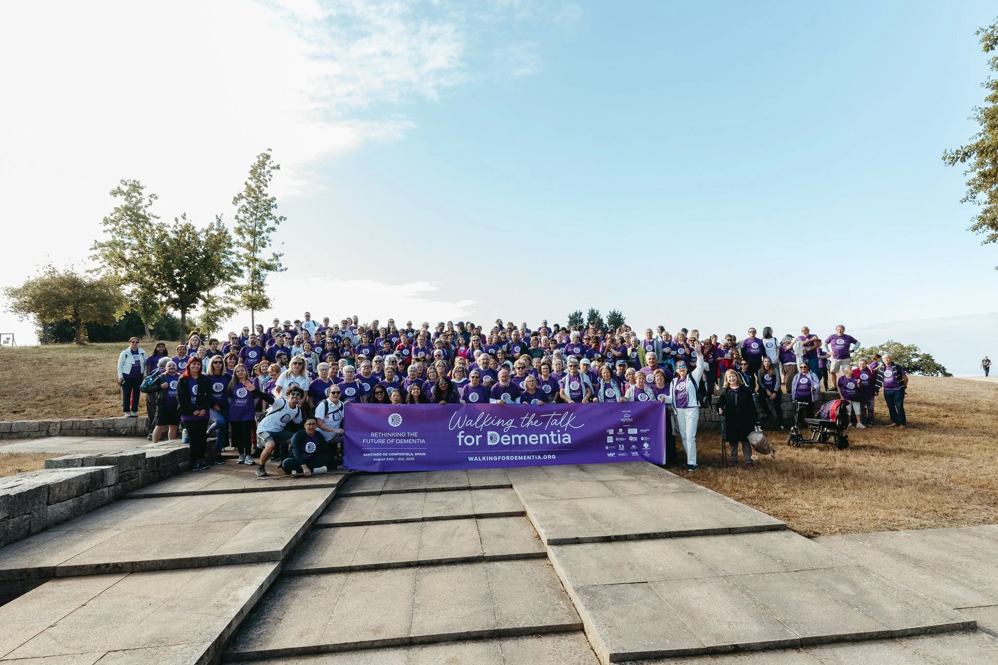 Group of people participating in a dementia awareness walk holding a purple banner that reads, 'Walking the talk for dementia, Rethinking the future of dementia.' The event takes place outdoors on a sunny day with trees in the background.