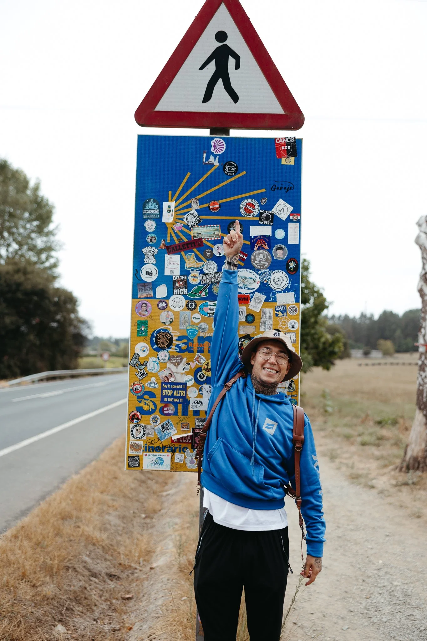 A smiling man in a blue hoodie, wearing glasses and a hat, pointing up at a collage-covered signpost next to a road.