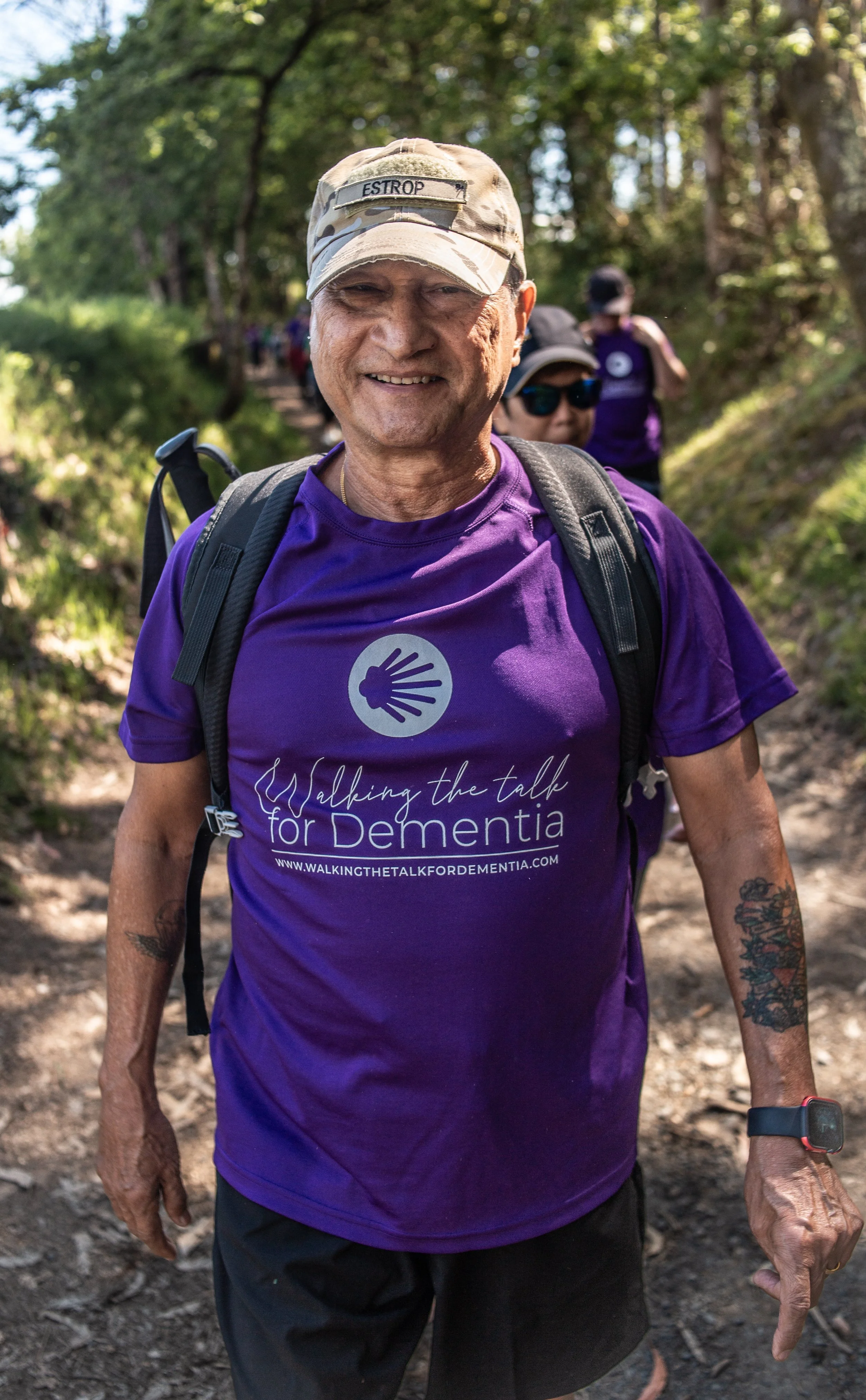 Smiling man wearing a purple 'Walking the Talk for Dementia' T-shirt, camouflage hat, and backpack walking on a forest trail with other hikers in the background.