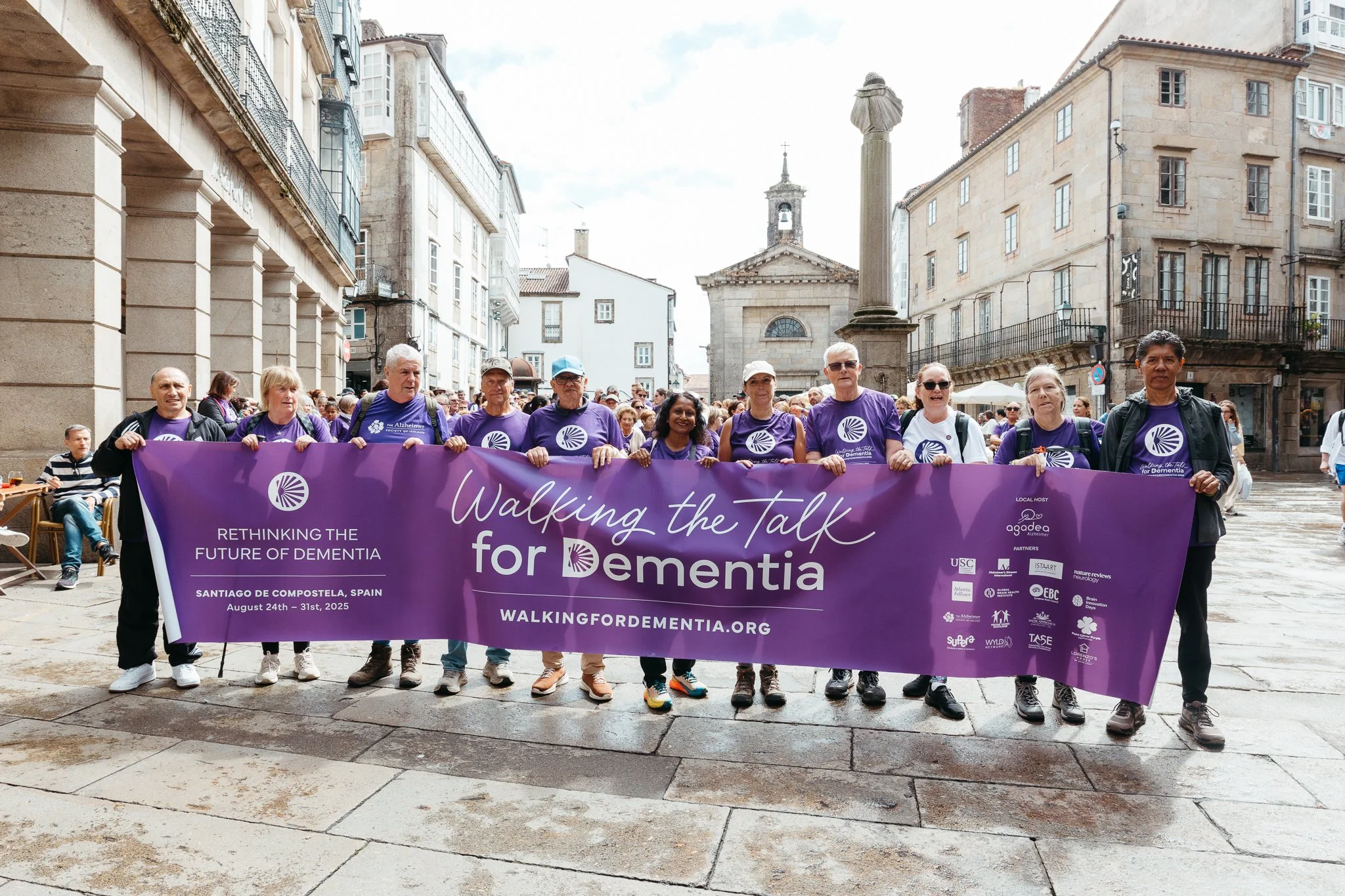 Group of people participating in a dementia awareness walk holding a large purple banner that reads "Walking the Talk for Dementia," in a historic city square with old buildings and a church in the background.