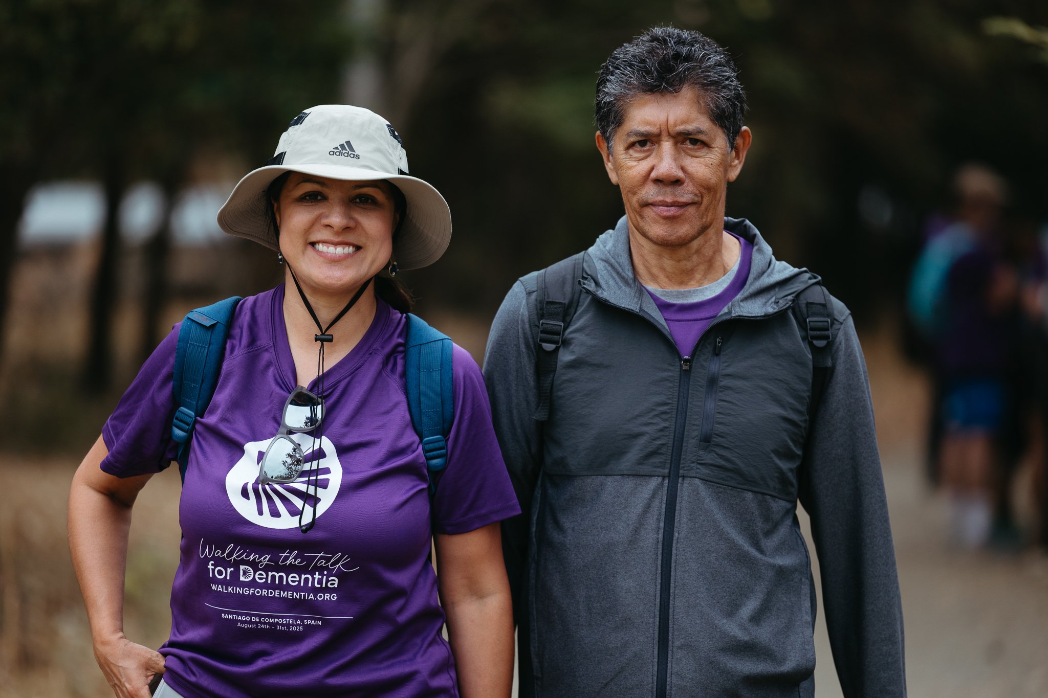 Two people, a woman and a man, standing outdoors in a wooded area. The woman is smiling, wearing a purple shirt with text about a dementia walking event, a white sun hat, and carrying a blue backpack. The man has a neutral expression, wearing a dark 