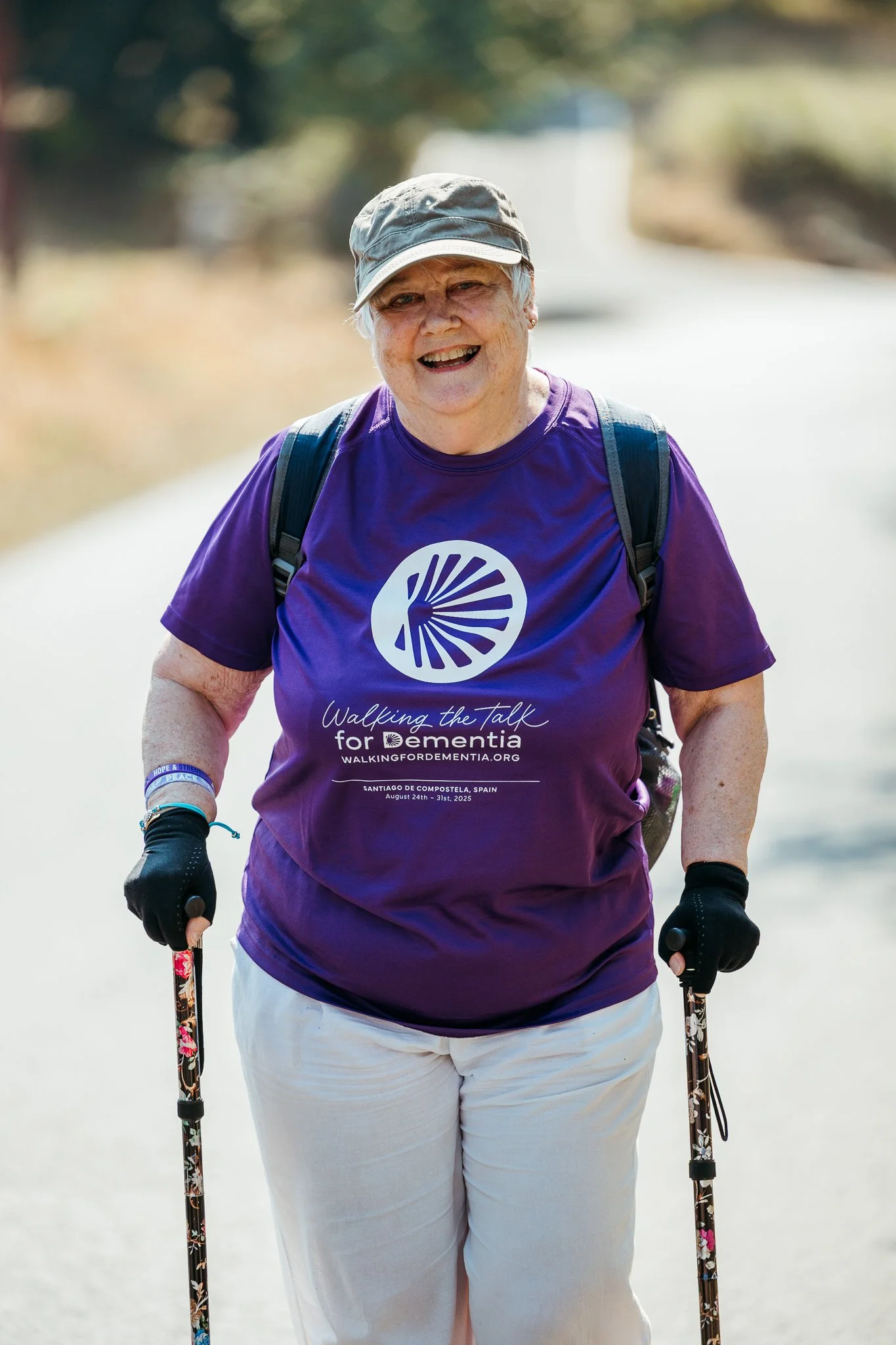 A smiling elderly woman participating in a walking event, wearing a purple t-shirt with white text and a logo, shorts, gloves, and holding walking poles on a sunny outdoor path.