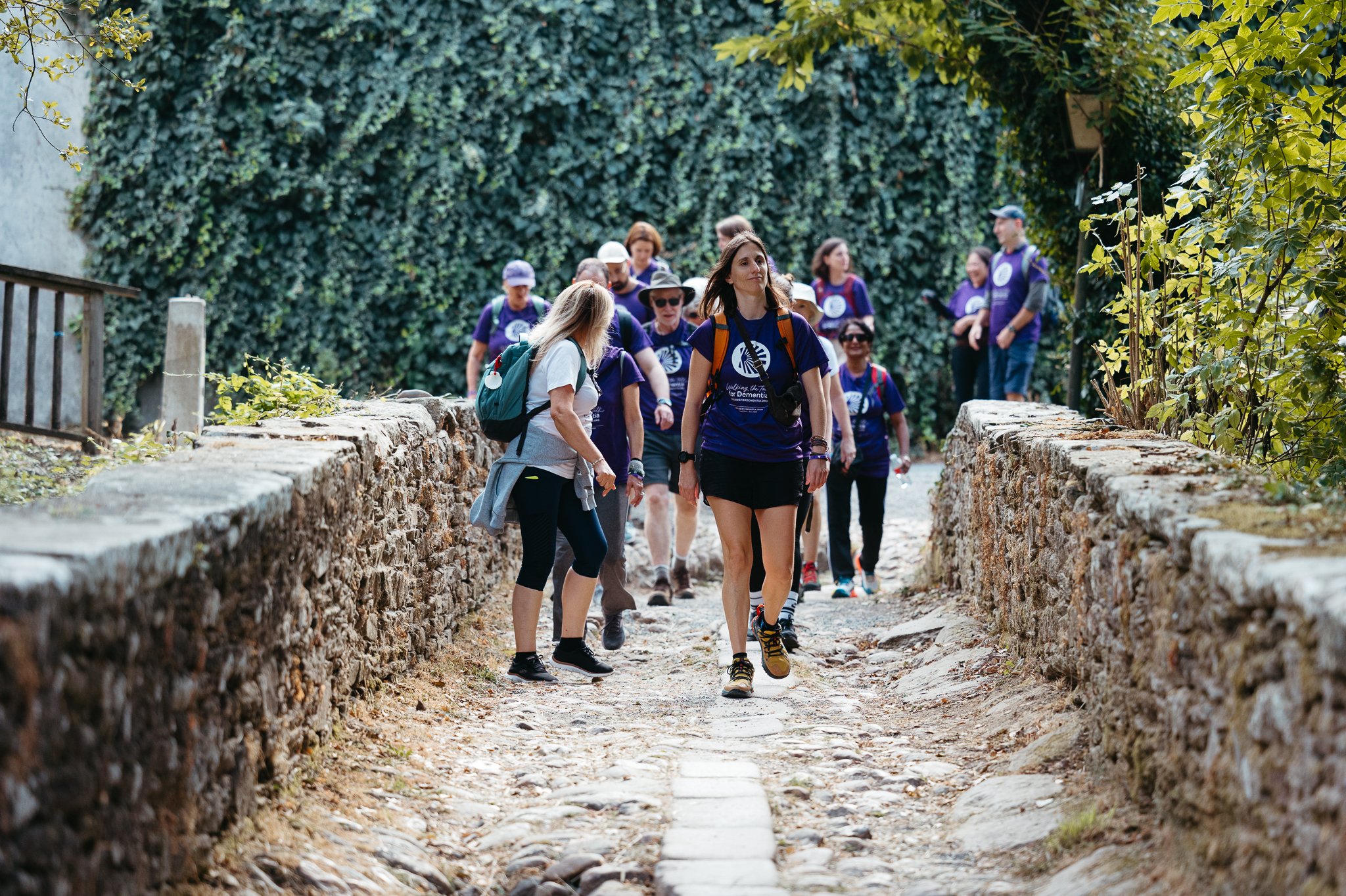 Group of people walking on a stone pathway surrounded by green foliage, some wearing hikers’ gear and purple shirts.